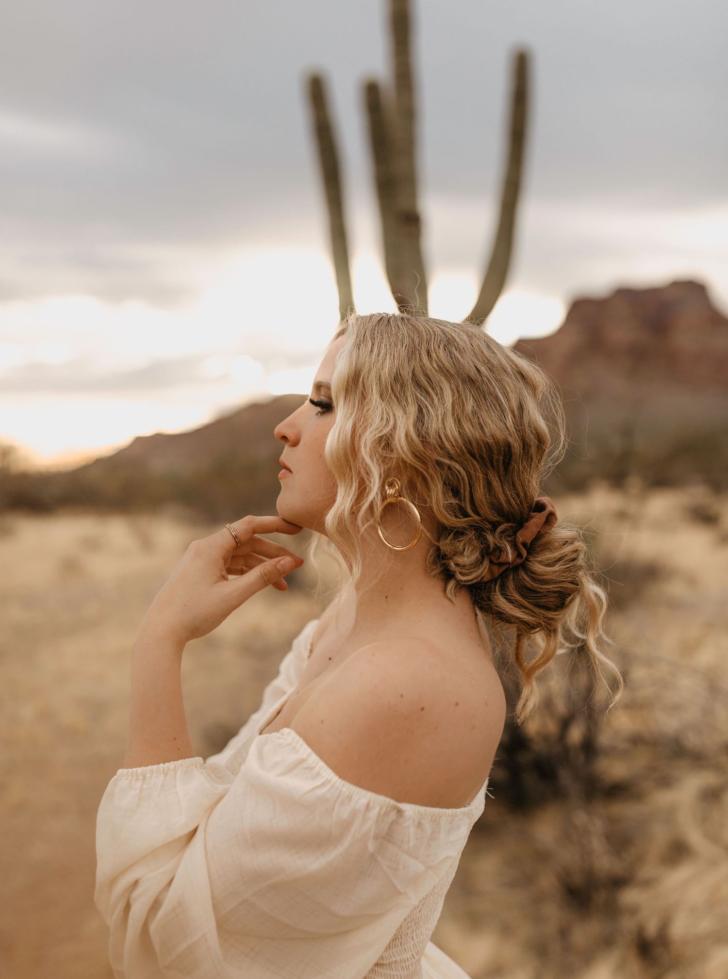 Ethereal desert portrait of woman in flowy white dress gazing up at sunset with cactus and mountains in background.