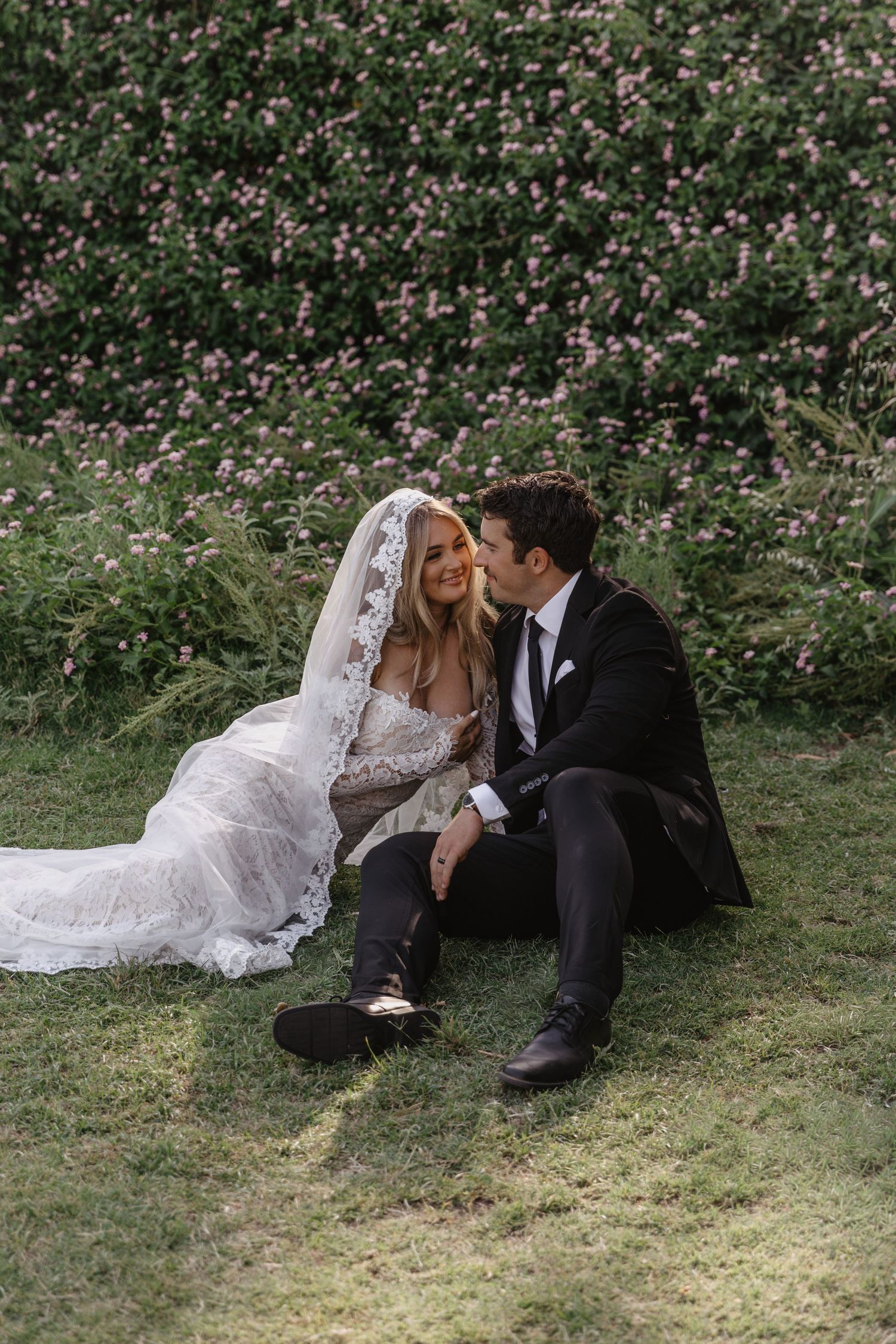 Bride and groom sitting on grass in front of flowering bushes, sharing an intimate moment on their wedding day.