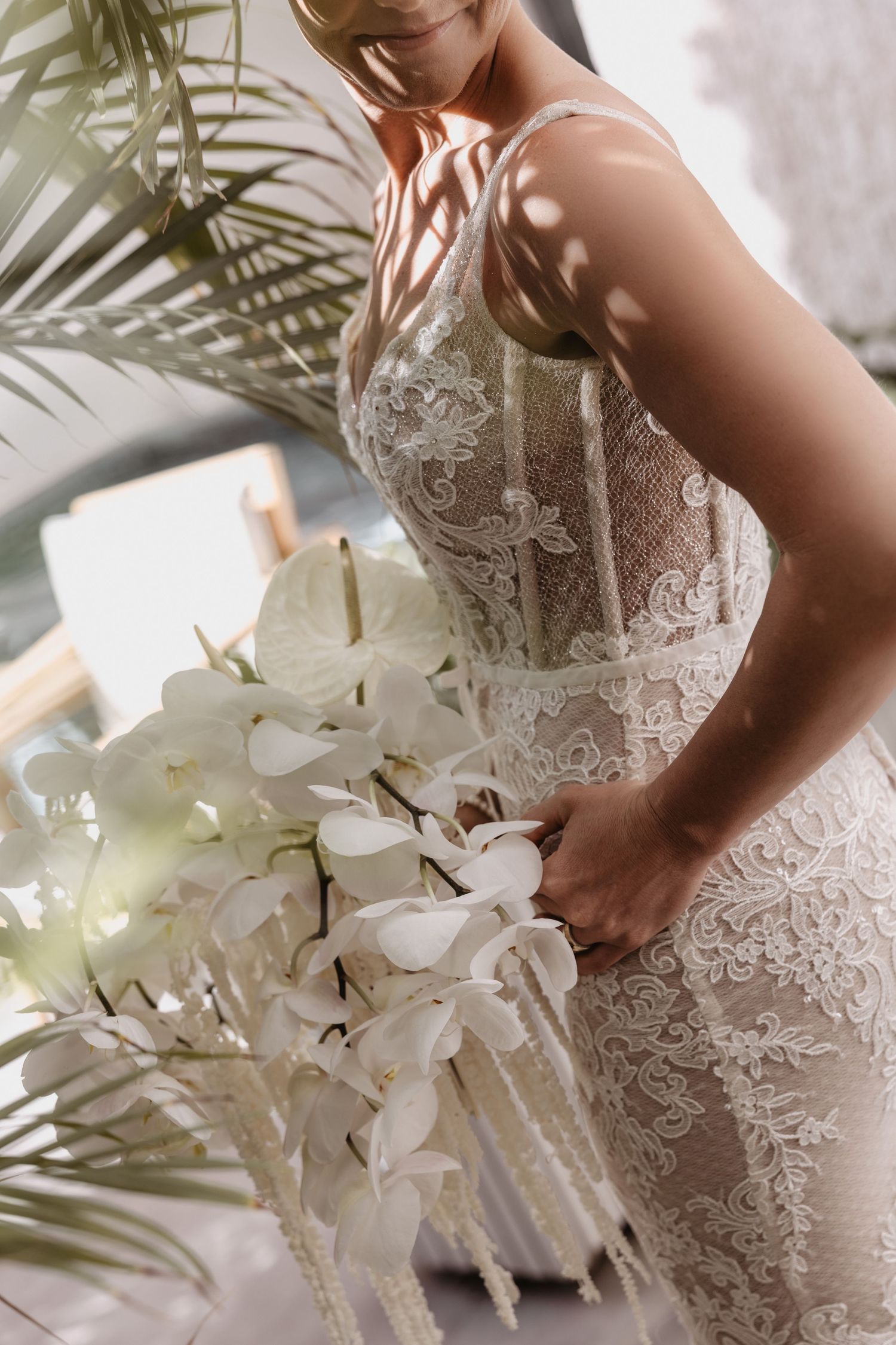 Elegant white lace wedding dress detail with cascading orchid bouquet and palm leaves in soft natural lighting.