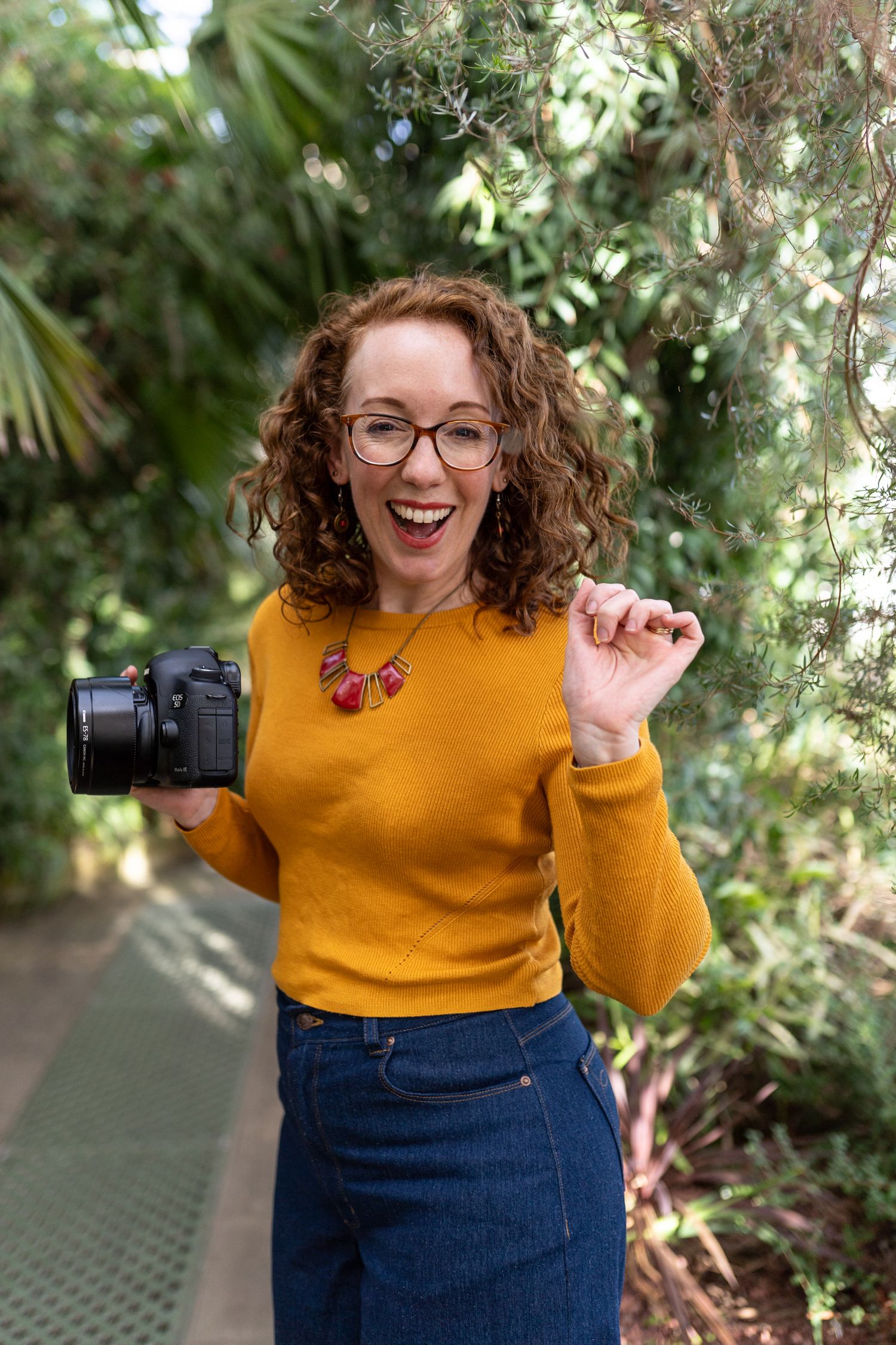 An enthusiastic photographer in yellow sweater holds camera among tropical foliage.