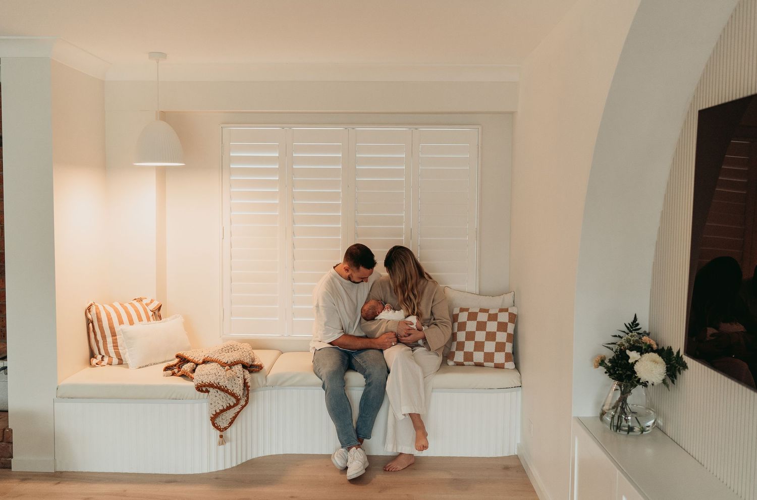 Two people sit close together on a cozy white window bench with decorative pillows in a warmly lit room.