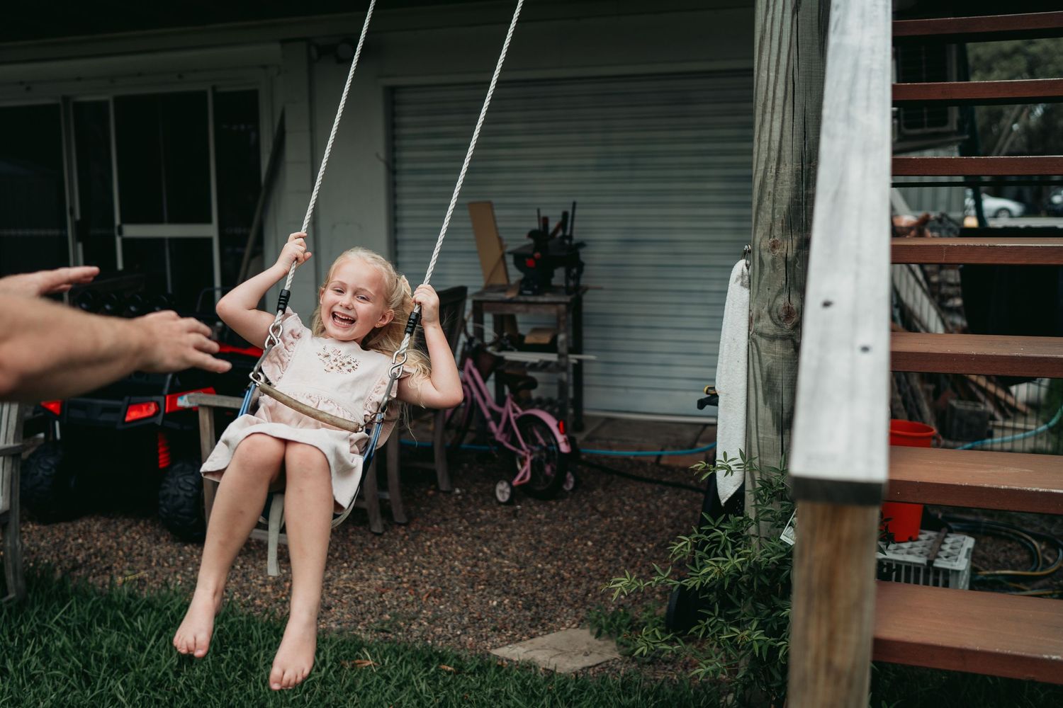 A young child enjoys swinging on a backyard rope swing while someone pushes them on a summer day.