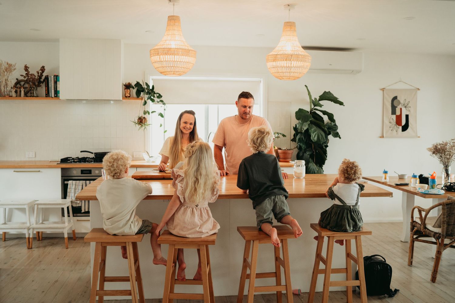 Family members sit at a wooden breakfast bar under crystal pendant lights in a bright modern kitchen interior.