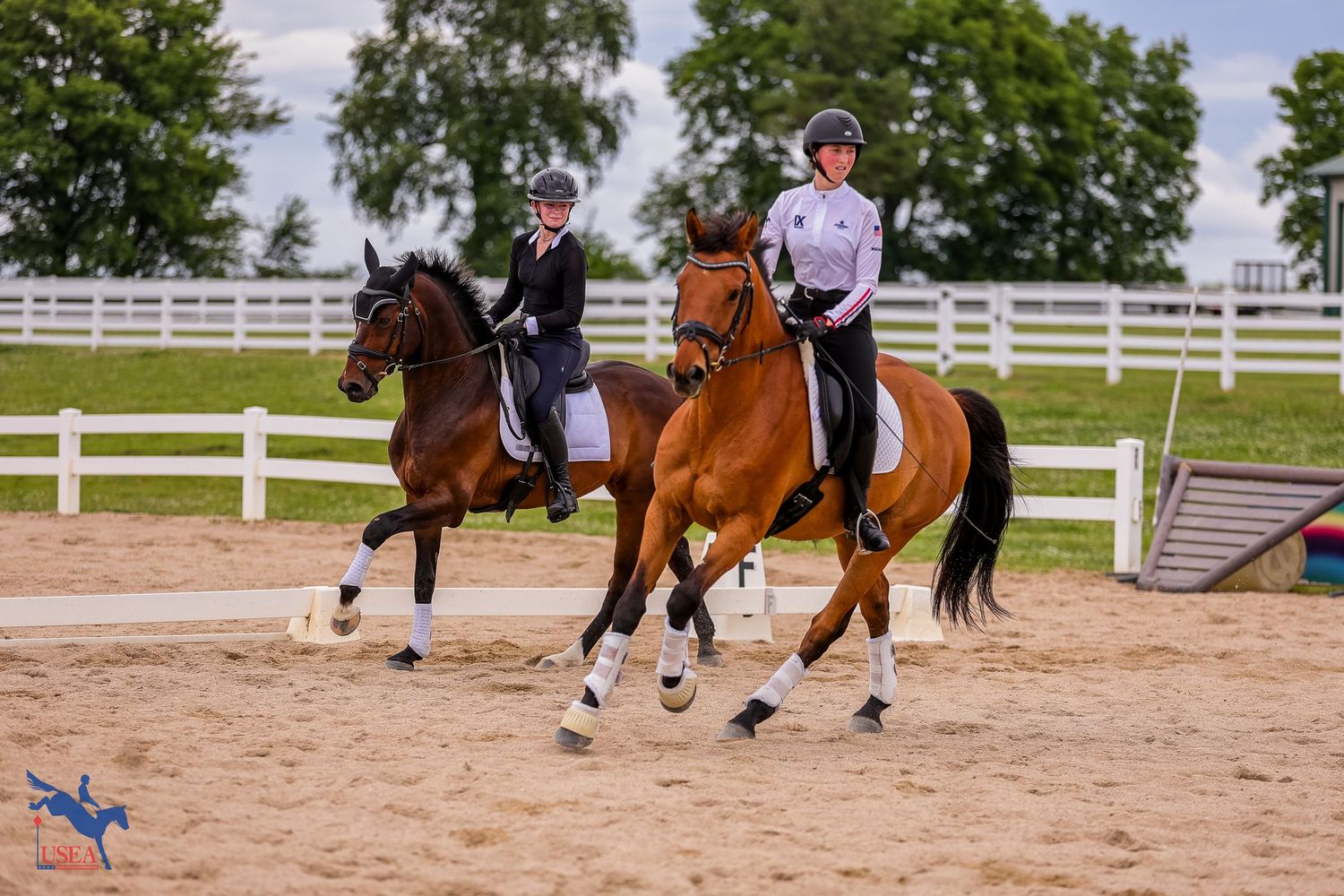 Two riders practicing dressage movements on horseback in a sand arena surrounded by white fencing.