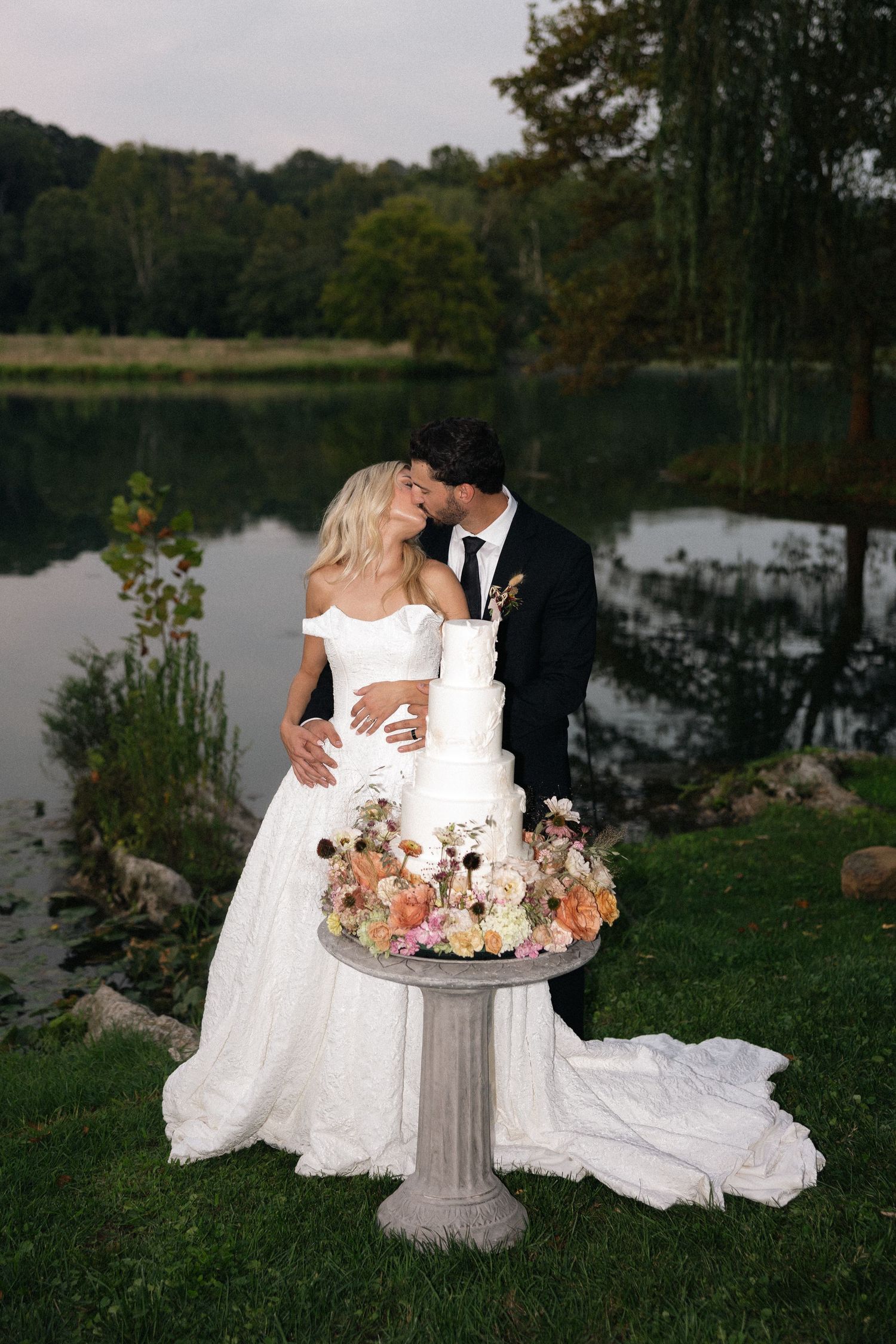 A romantic wedding couple poses with their elegant white tiered cake by a serene lake.