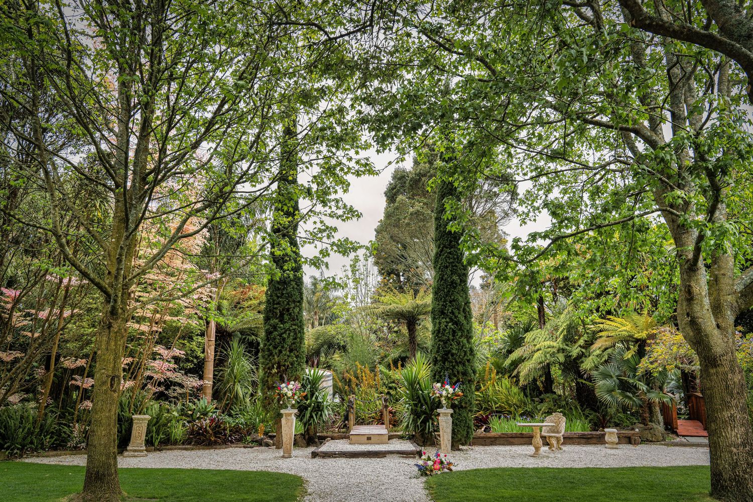Lush garden with tall cypress trees, green lawn, gravel path, and ornamental statue surrounded by vibrant flowering plants.