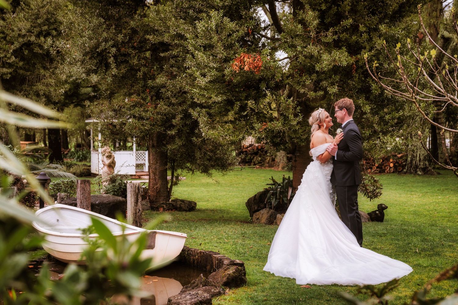 Bride and groom share a romantic moment in a lush garden setting with a rustic boat and trees at their wedding.