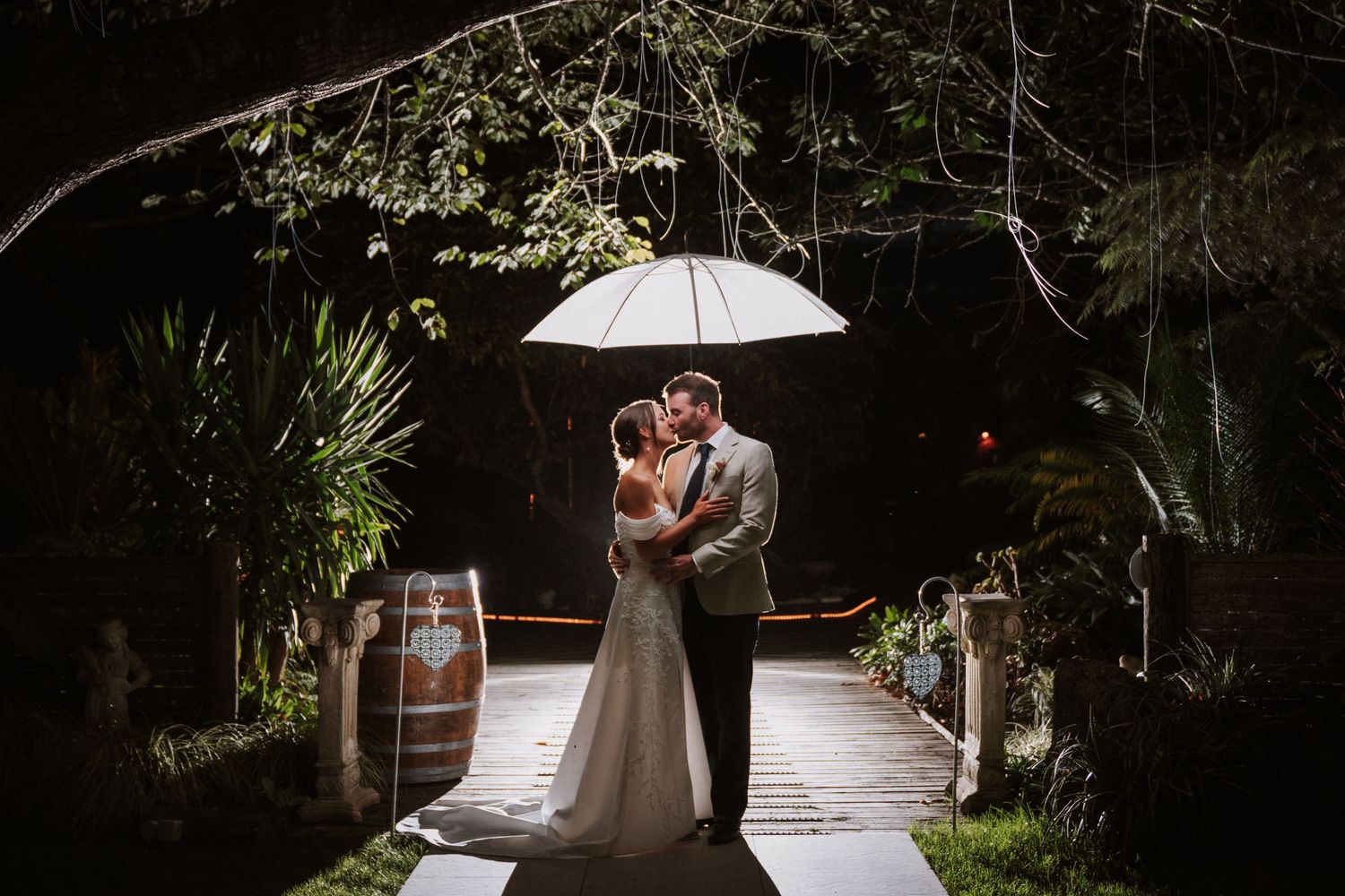 Bride and groom kissing under a white umbrella on a romantic rainy night at an outdoor wedding venue.