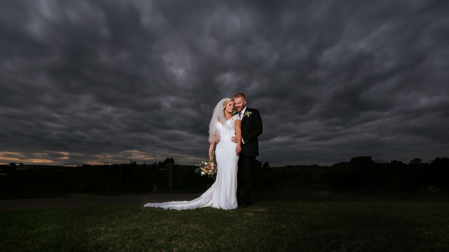 Bride and groom embrace under dramatic stormy dark clouds at dusk on a green lawn in an elegant outdoor wedding portrait.