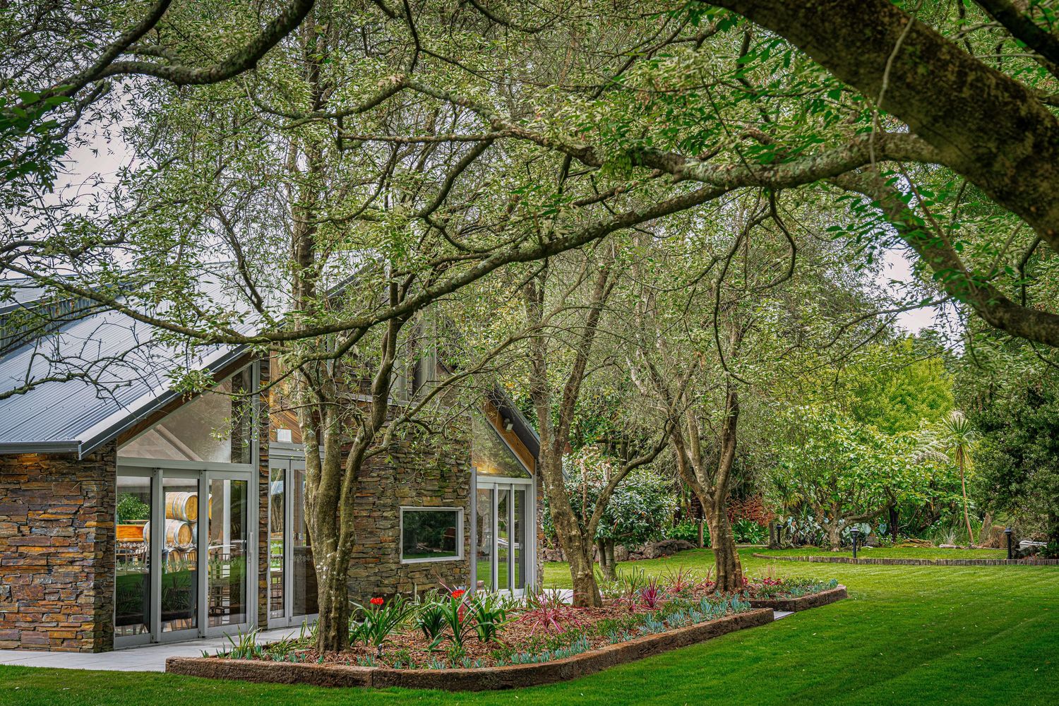 Stone cottage surrounded by tall trees and lush green lawn with colorful flower beds in a serene garden setting.