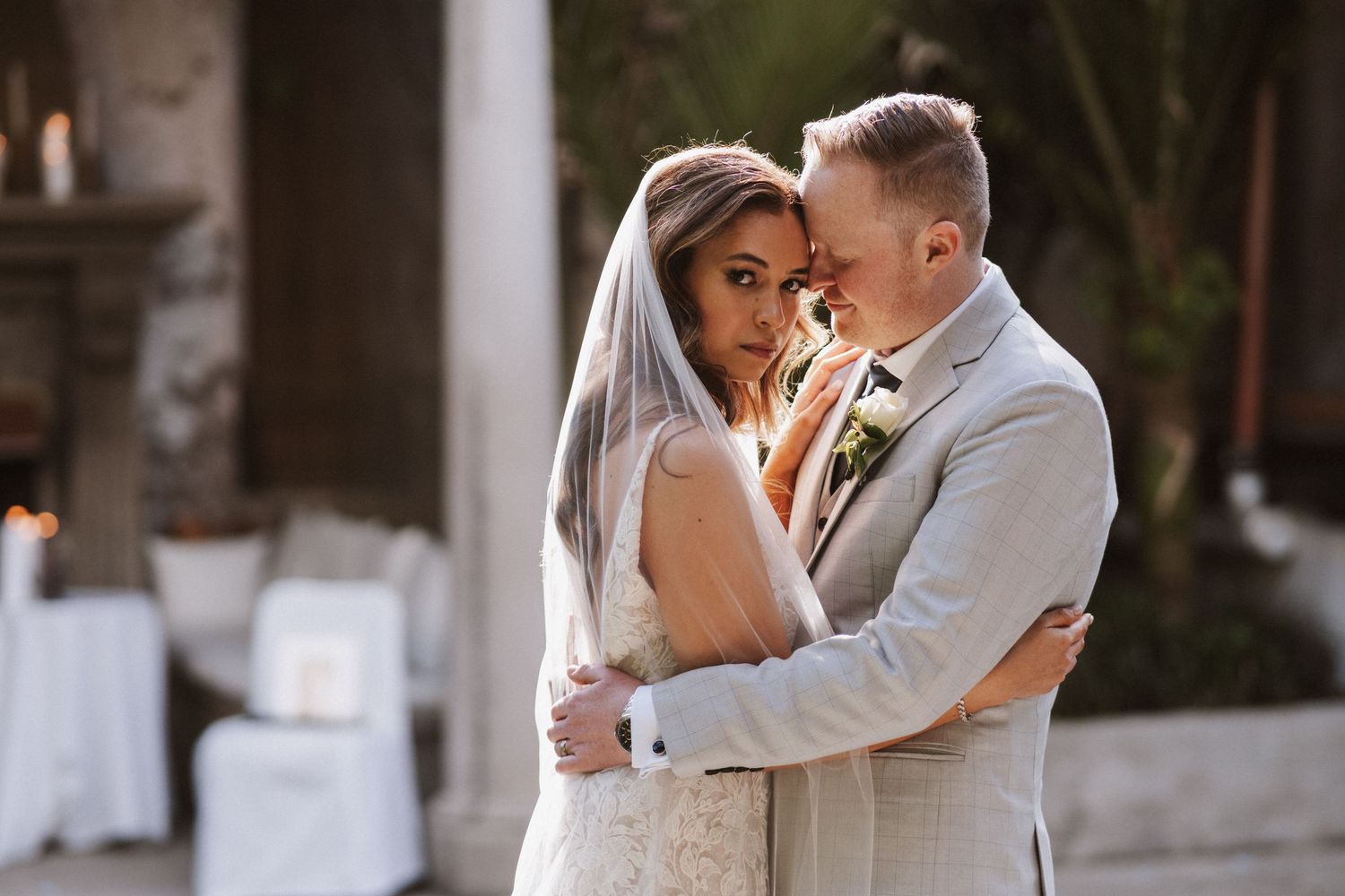 Bride and groom embrace outdoors at elegant wedding venue, bride in lace gown with veil, groom in light gray suit.