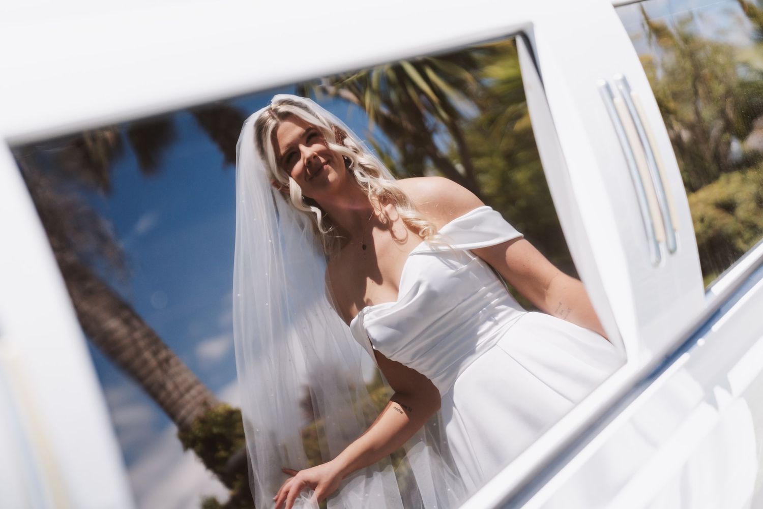 Bride in white off-shoulder gown and veil reflected in a car window, with palm trees in the background.