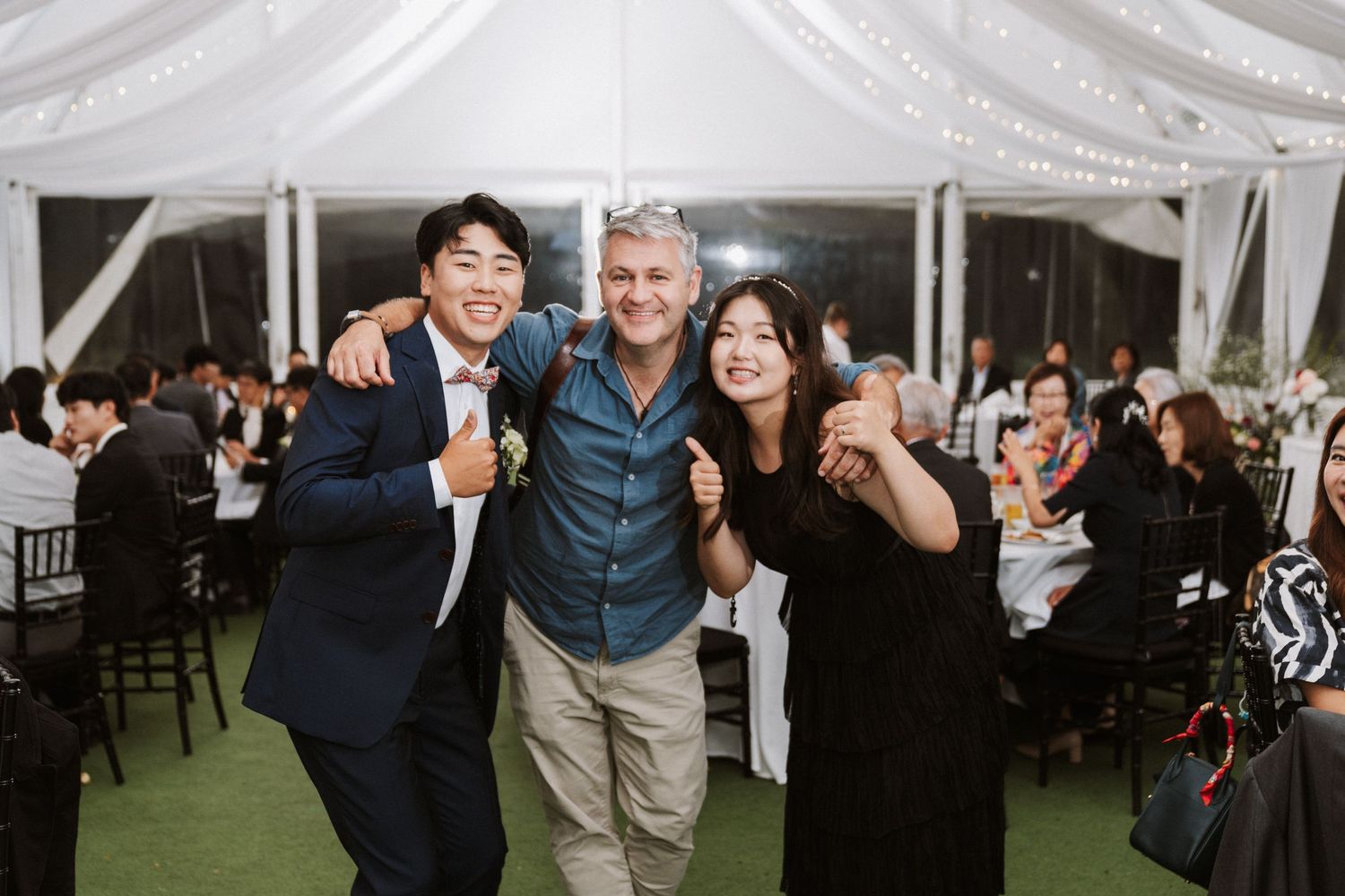 Three guests smile and pose together at an elegant outdoor wedding reception under a white tent with string lights.