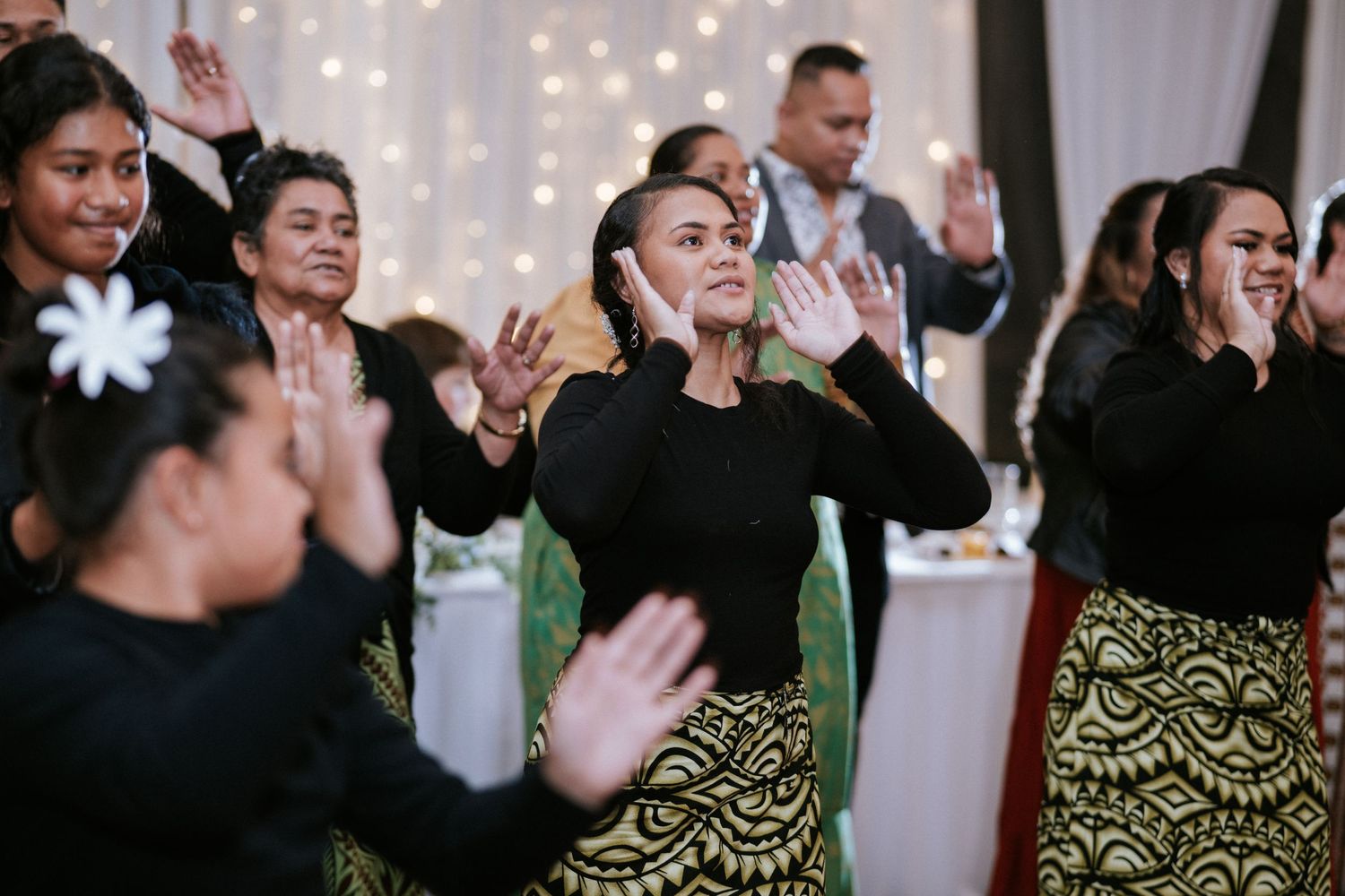 Women in traditional Polynesian skirts perform a cultural dance at a festive indoor celebration with fairy lights.