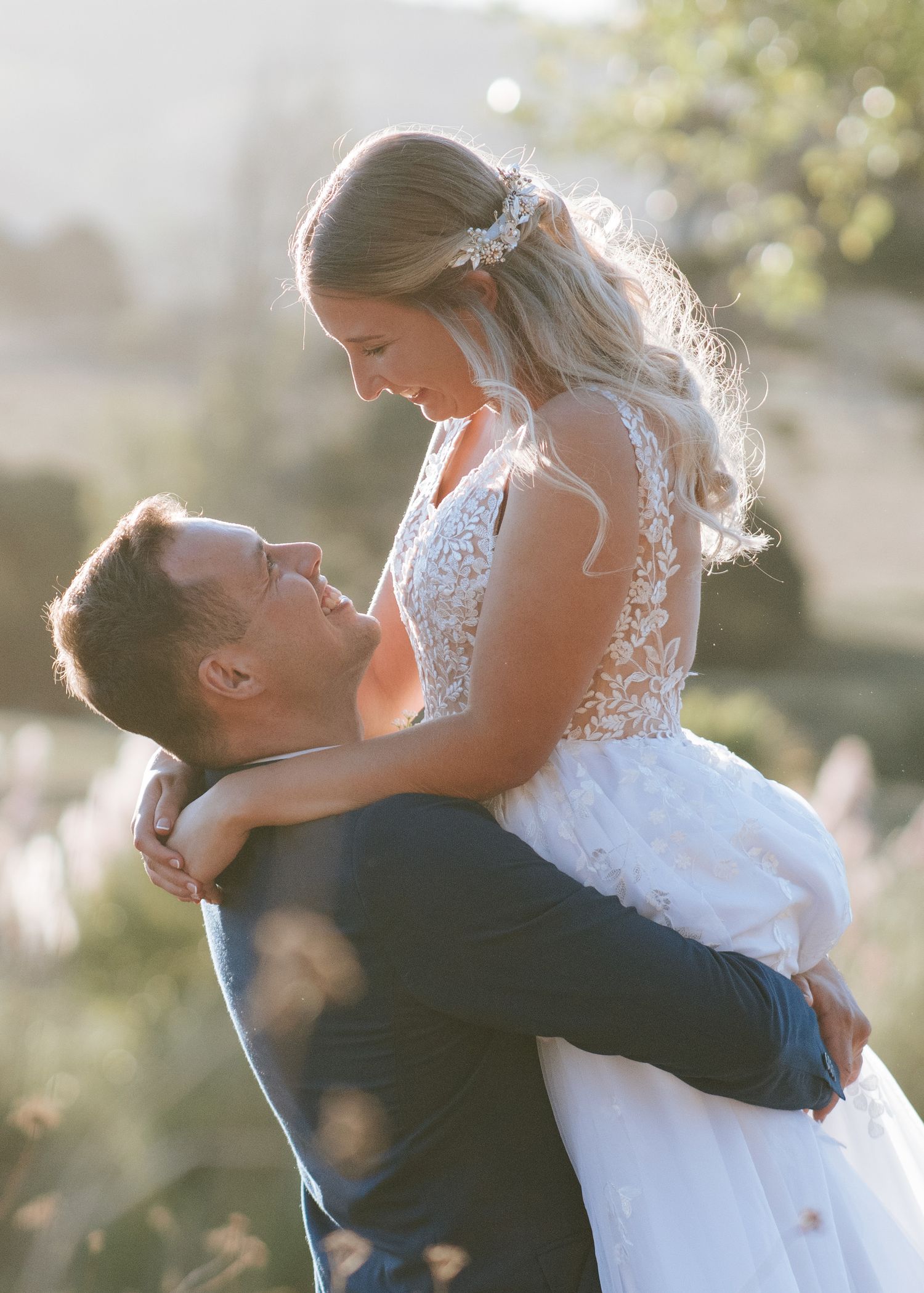 Groom lifting bride in lace wedding dress outdoors in golden sunlight, sharing a romantic moment.