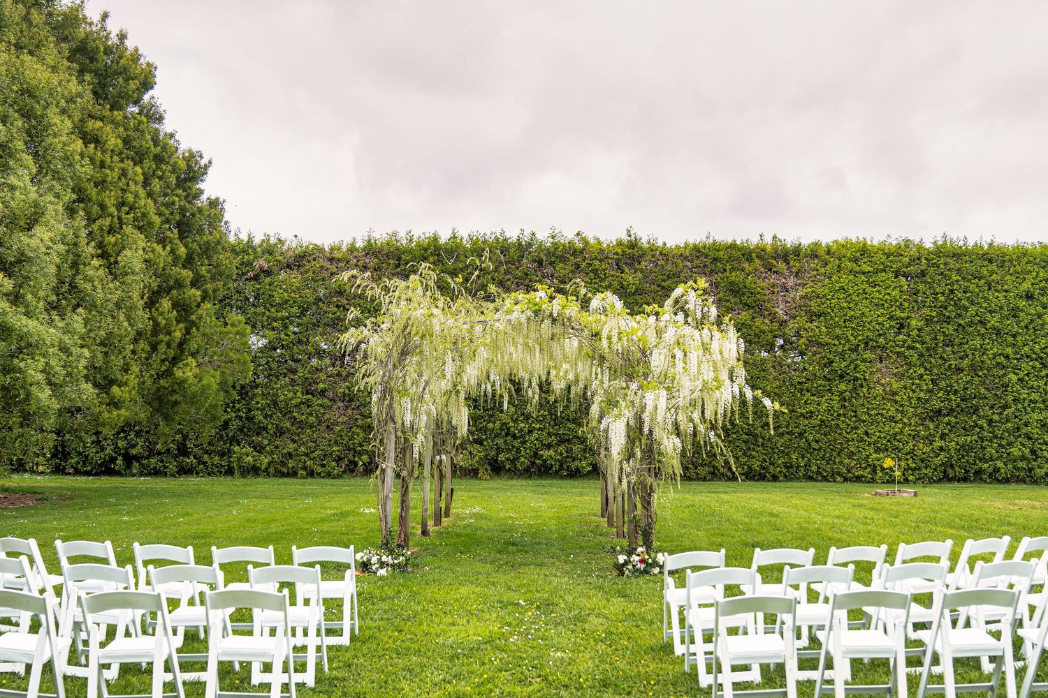 Outdoor wedding ceremony setup with white chairs on green lawn, floral arch, and lush hedge backdrop.