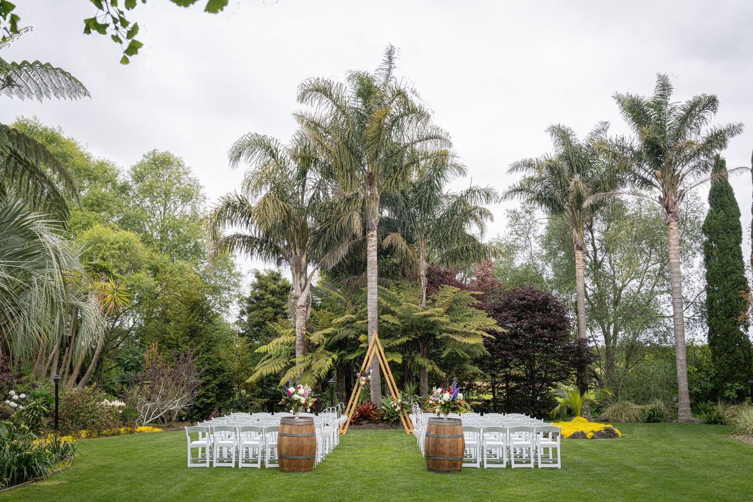 Outdoor wedding ceremony setup with white chairs, barrel arch, and lush palm trees on manicured lawn.
