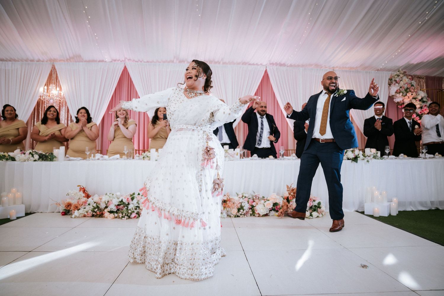 Bride in white lace gown dancing with groom in navy suit at elegant wedding reception with pink floral decor.