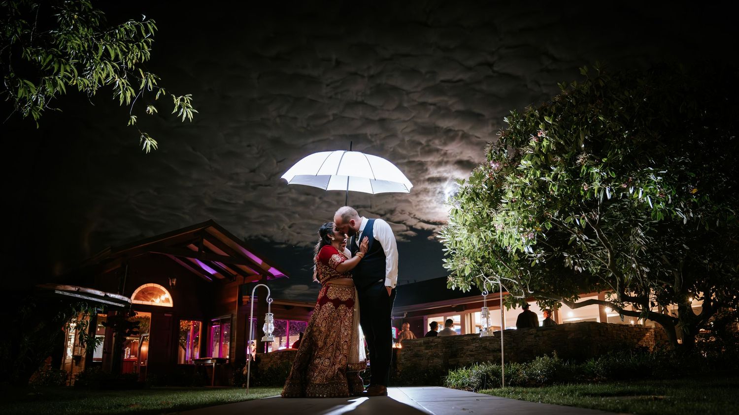 Couple sharing a romantic kiss under a white umbrella at night with dramatic cloudy sky and warmly lit venue.