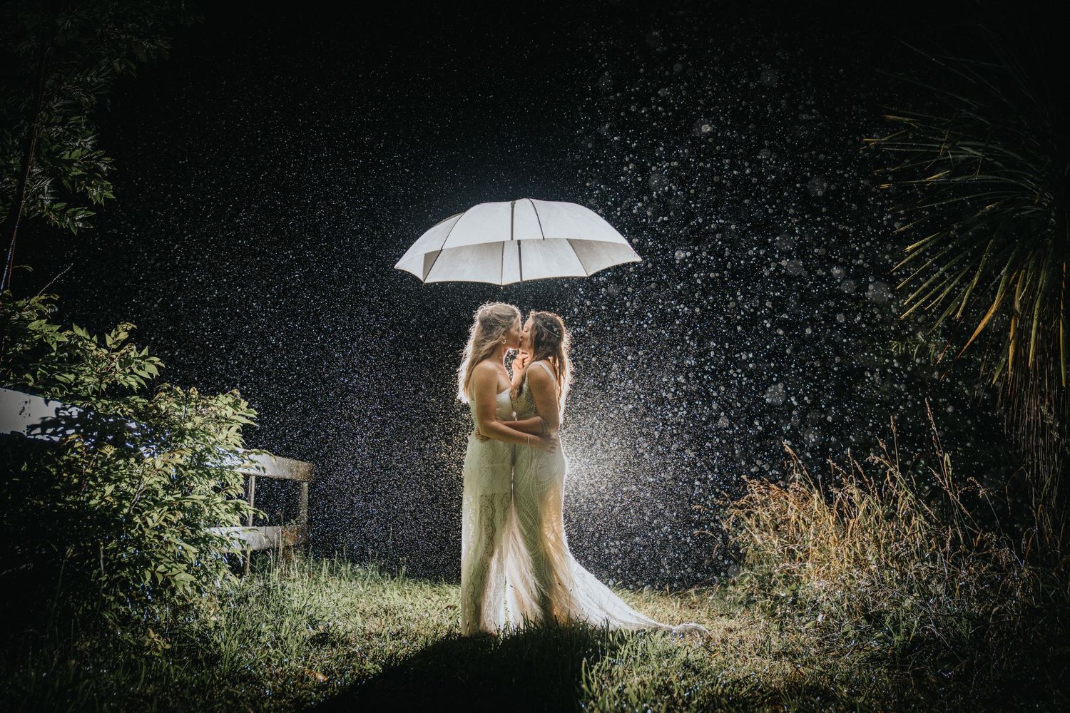 Two brides sharing a romantic kiss under a white umbrella in the rain at night during their wedding.