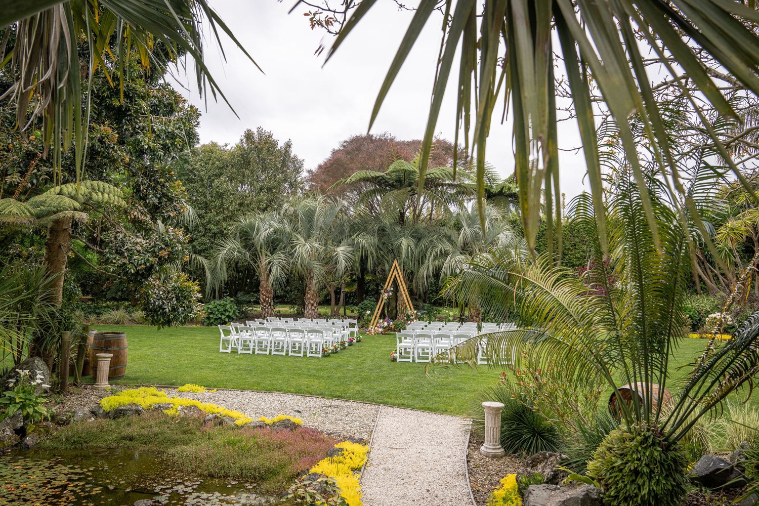 Lush garden wedding ceremony setup with white chairs and a wooden arch surrounded by tropical palms and greenery.