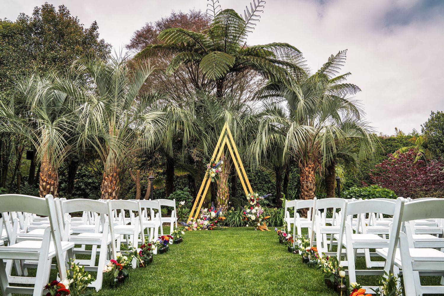 Outdoor wedding ceremony setup with white chairs, tropical palm trees, and a gold triangular arch adorned with colorful flowers.