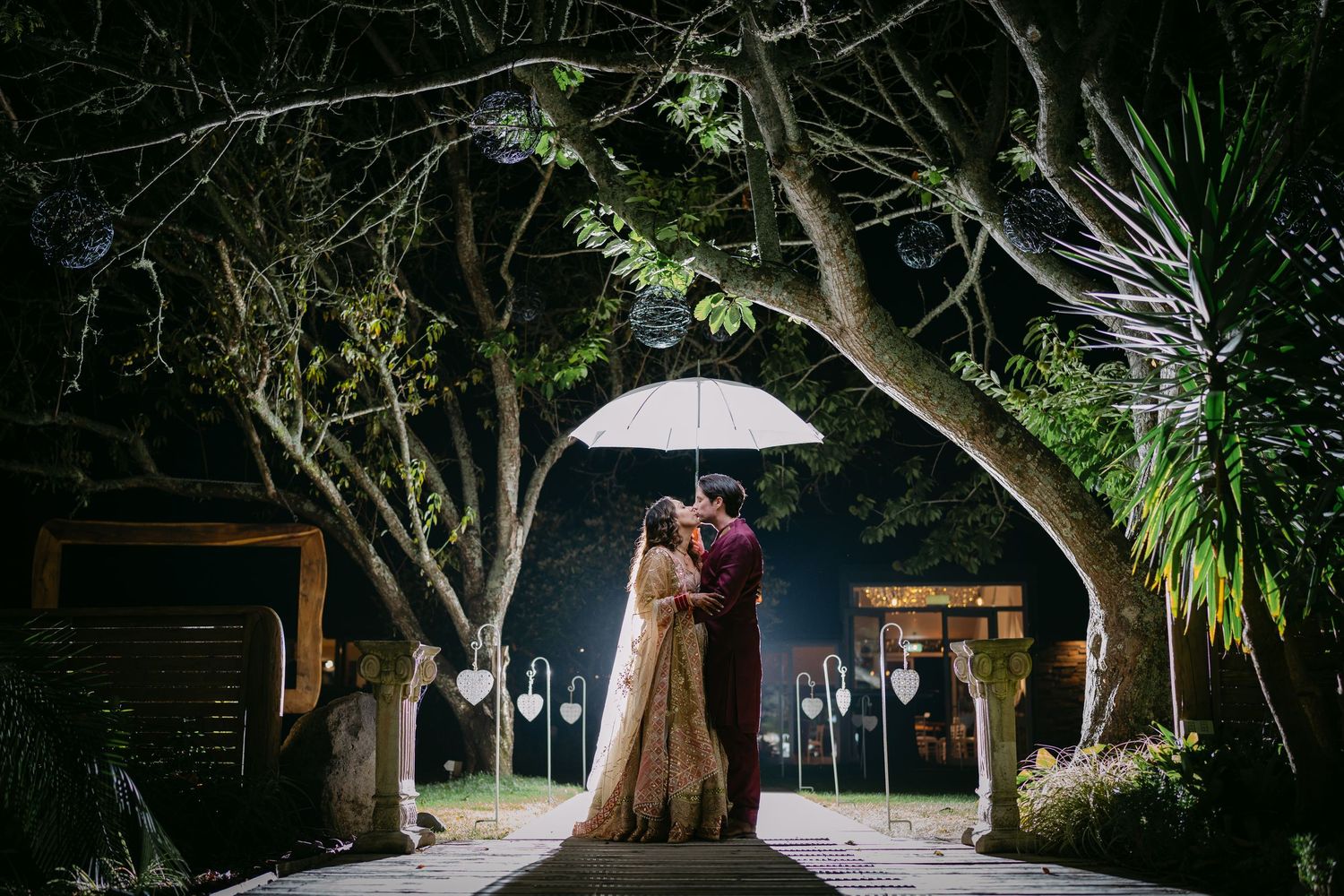 Couple sharing romantic moment under white umbrella on tree-lined path at night during Indian wedding celebration.