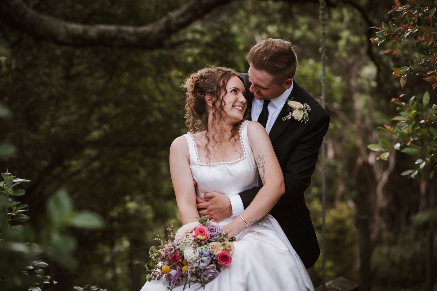A bride and groom share a romantic moment in a lush forest setting, with the bride holding a colorful bouquet.