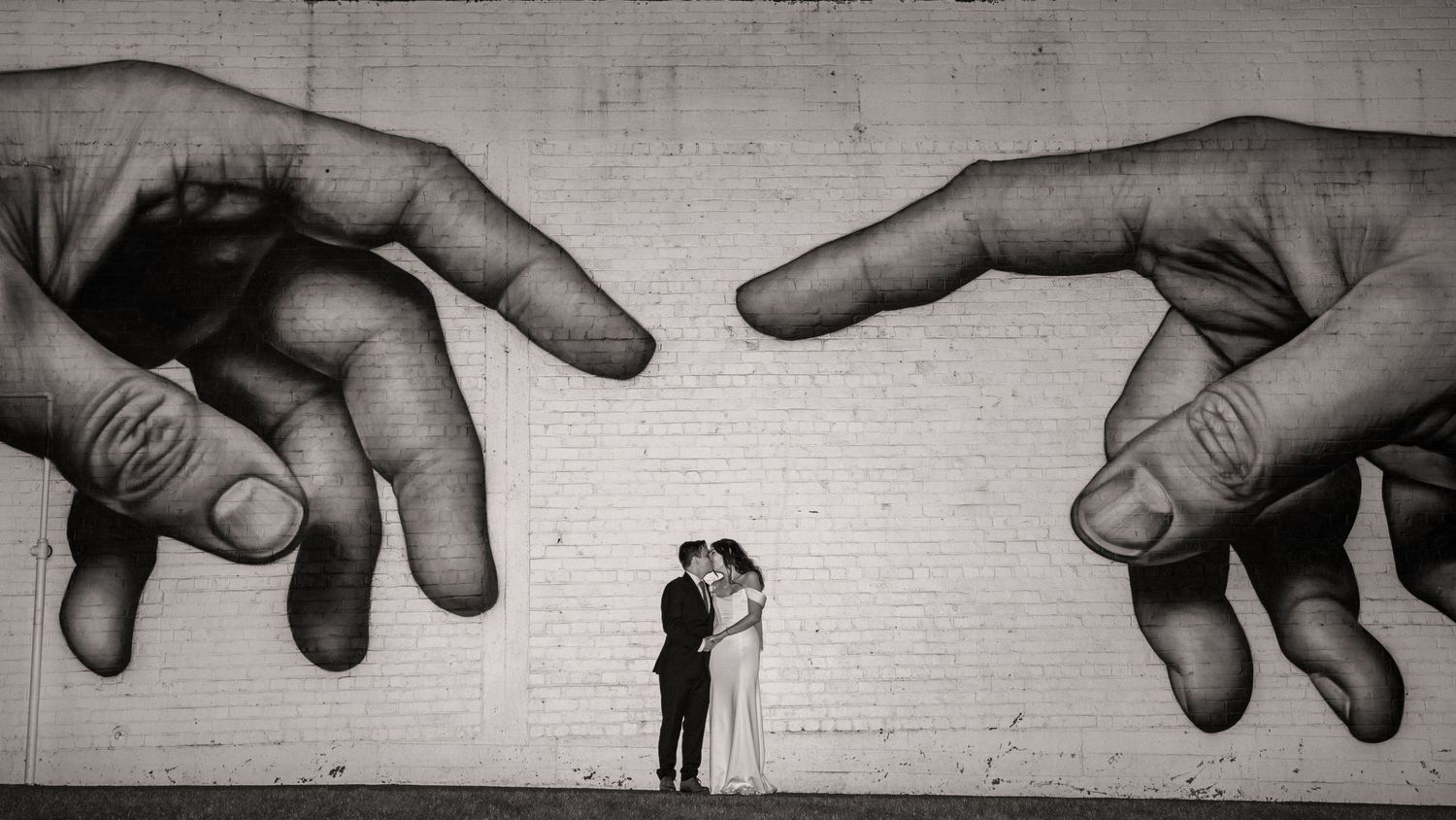 Couple embraces beneath giant black and white mural of two hands almost touching, inspired by Michelangelo's Creation of Adam.