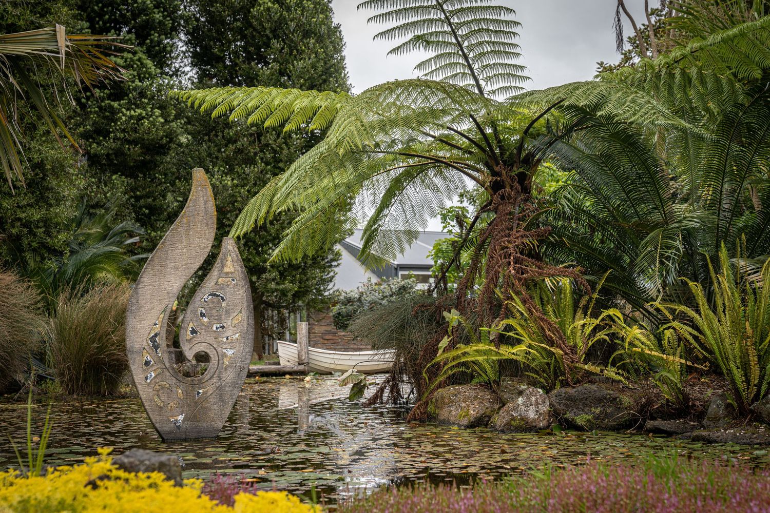 Stone sculpture beside a tranquil pond surrounded by lush ferns and tropical plants in a serene garden setting.