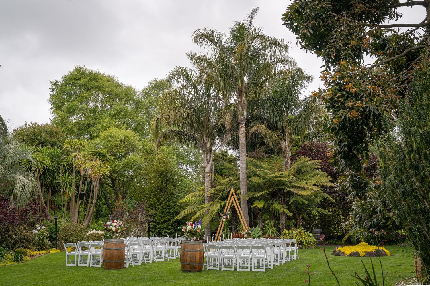 Outdoor wedding ceremony setup with white chairs and barrel decor surrounded by lush gardens and tall palm trees.