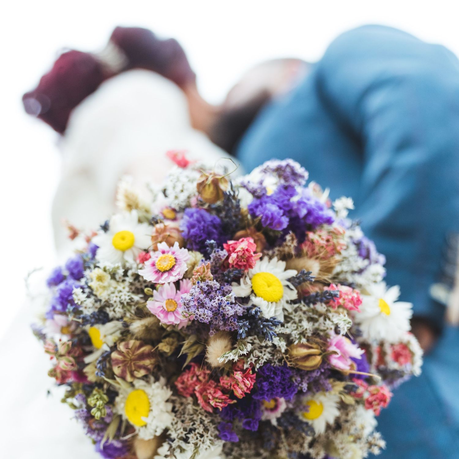 A couple shares a romantic kiss behind a vibrant wildflower bouquet with daisies, lavender, and colorful blooms.