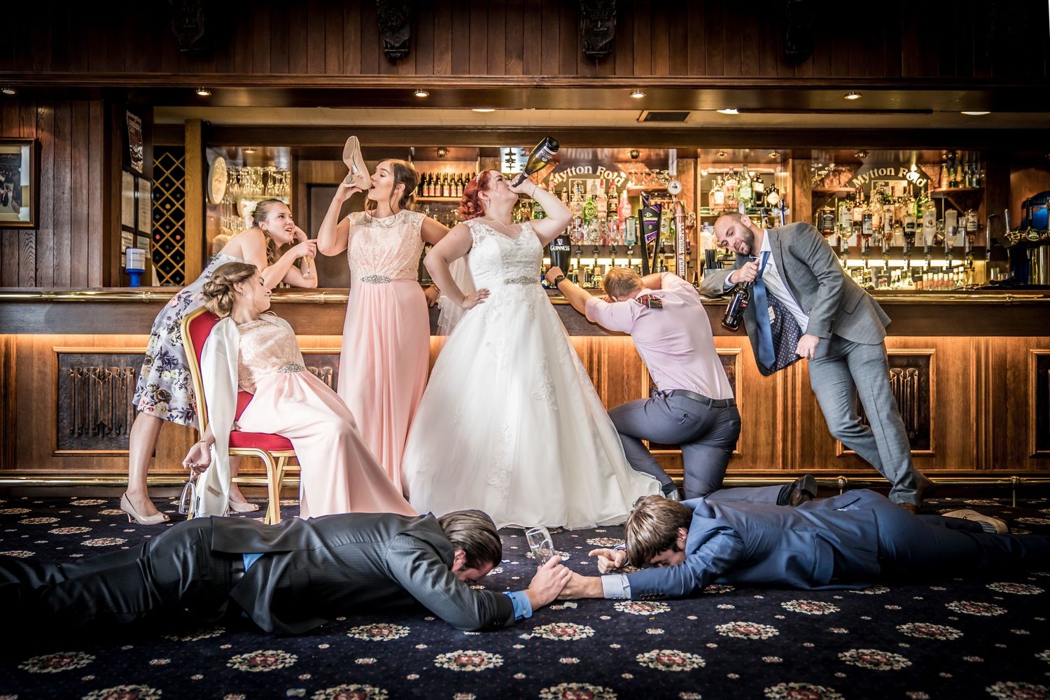 Wedding party poses dramatically at a bar, with groomsmen on floor and bridesmaids celebrating in a fun group photo.