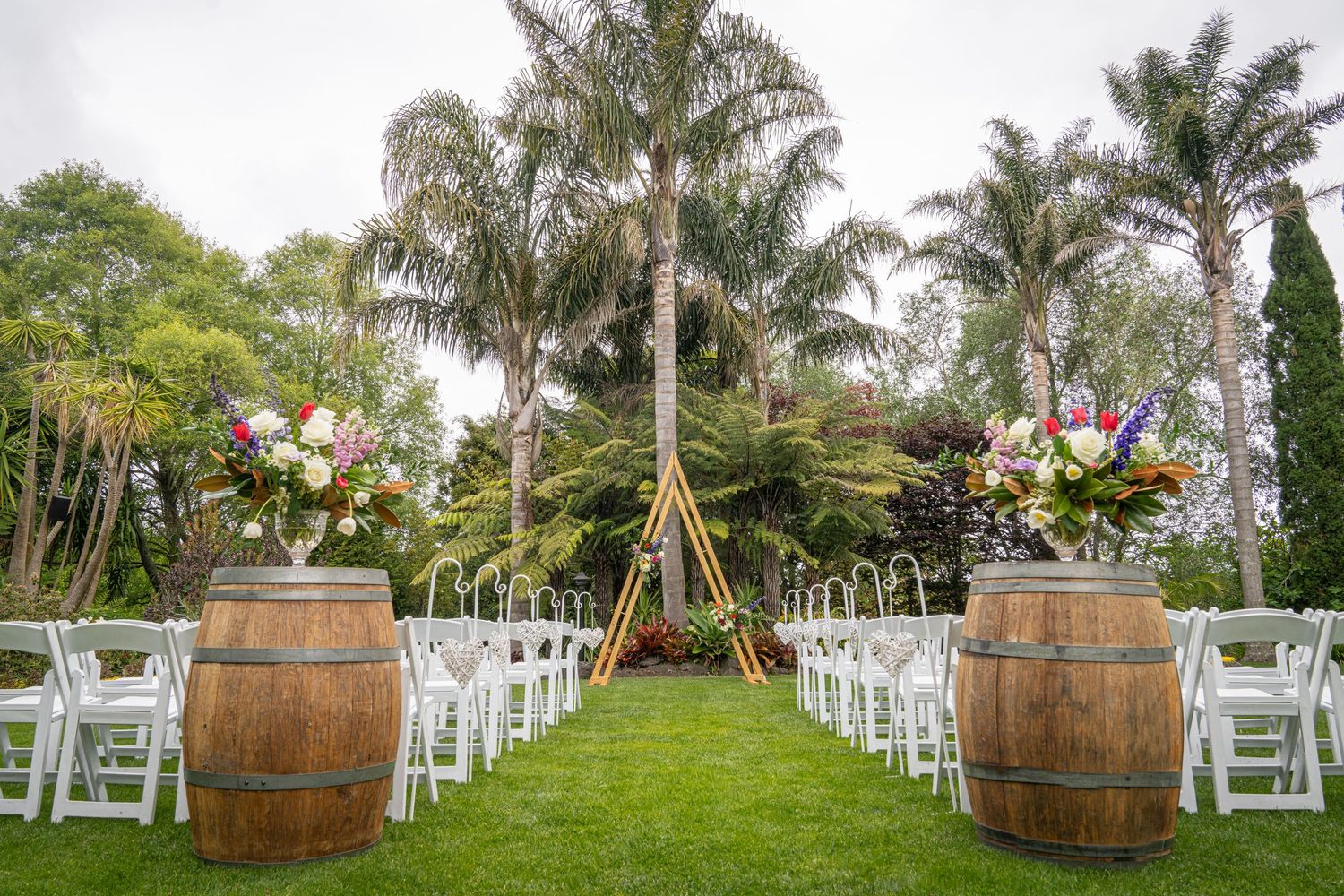 Outdoor wedding ceremony setup with white chairs, wooden barrels topped with floral arrangements, and palm trees in background.
