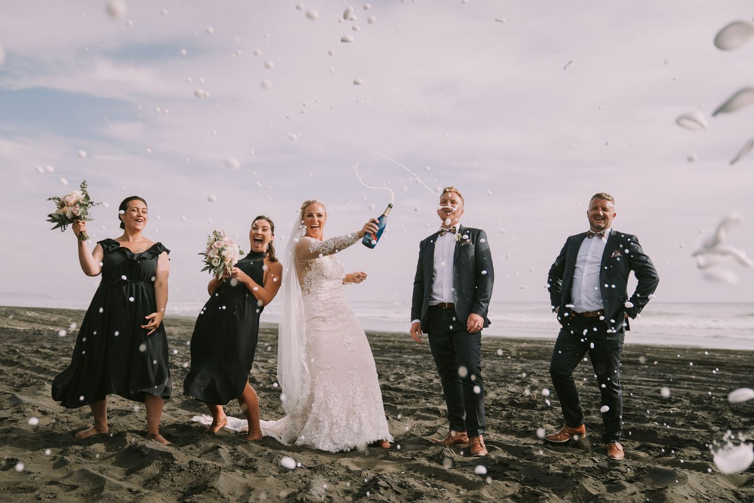 Wedding party celebrating on beach, bride popping champagne with bridesmaids in black dresses and groomsmen in navy suits.