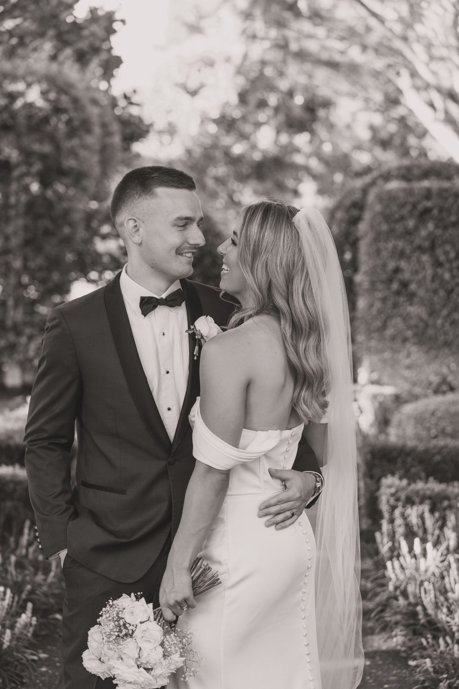 Black and white photo of a smiling bride in off-shoulder gown and groom in tuxedo sharing a romantic moment in a garden.
