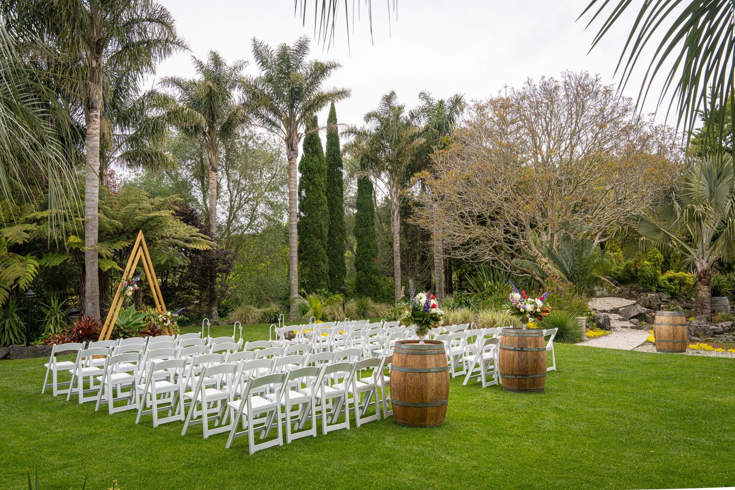 Outdoor wedding ceremony setup with white chairs, wooden barrels, and a triangular arch on a lush green lawn surrounded by palm trees.