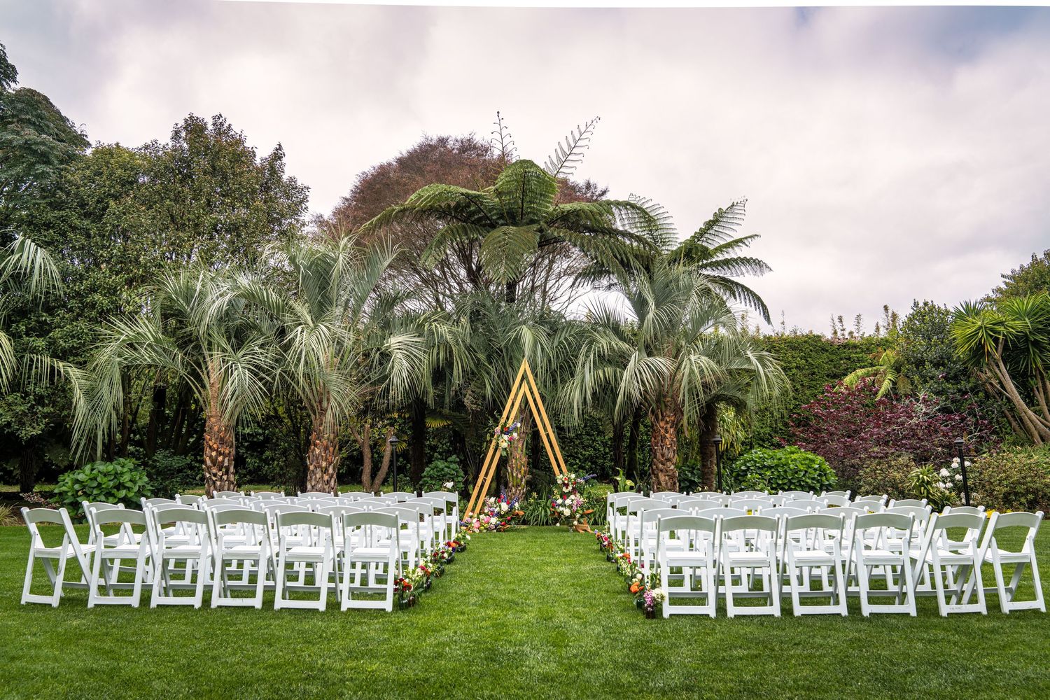Outdoor wedding ceremony setup with white chairs, golden triangular arch, and lush palm trees on green lawn.