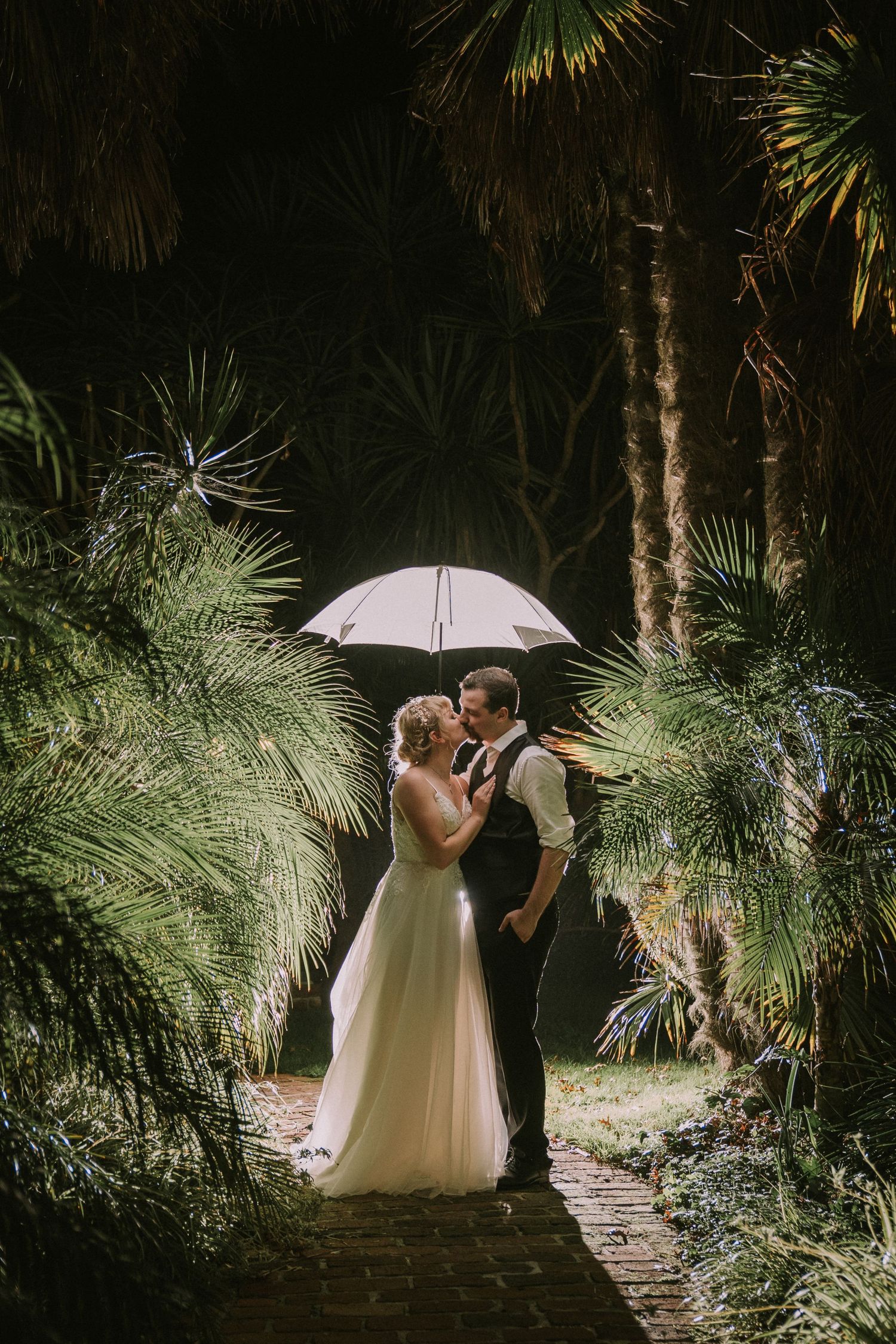 Bride and groom kissing under a glowing white umbrella surrounded by lush palm trees on a romantic rainy wedding night.