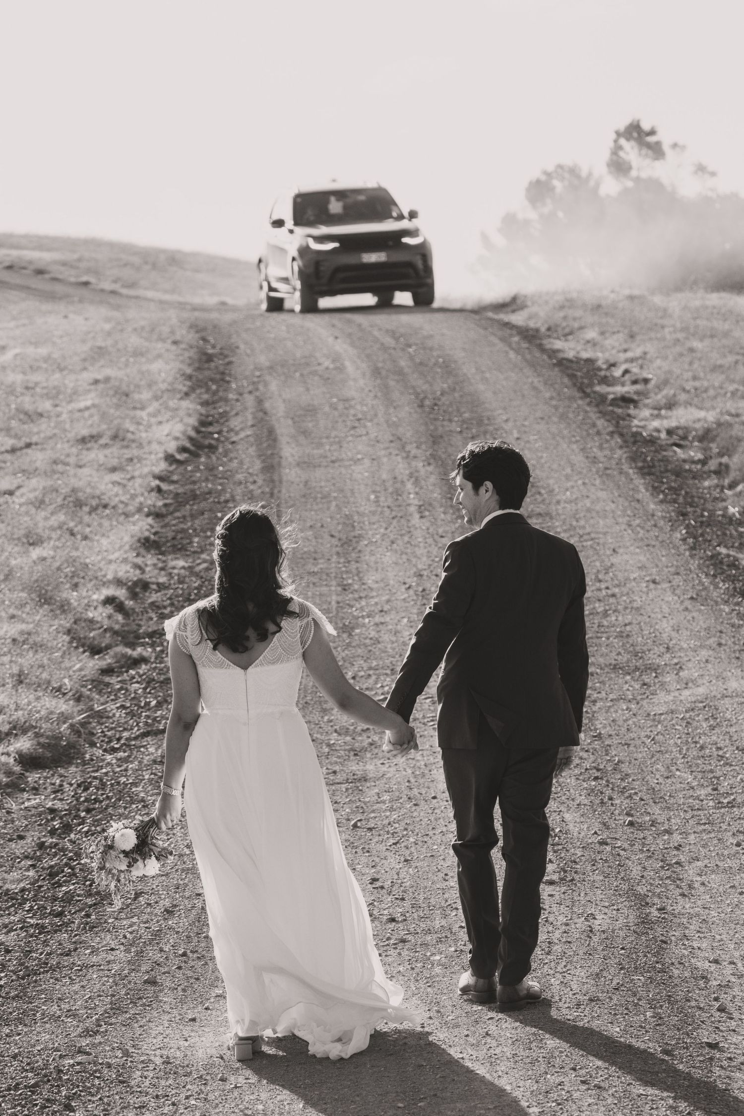 Black and white photo of bride and groom holding hands on a dirt road with a car approaching in the distance.