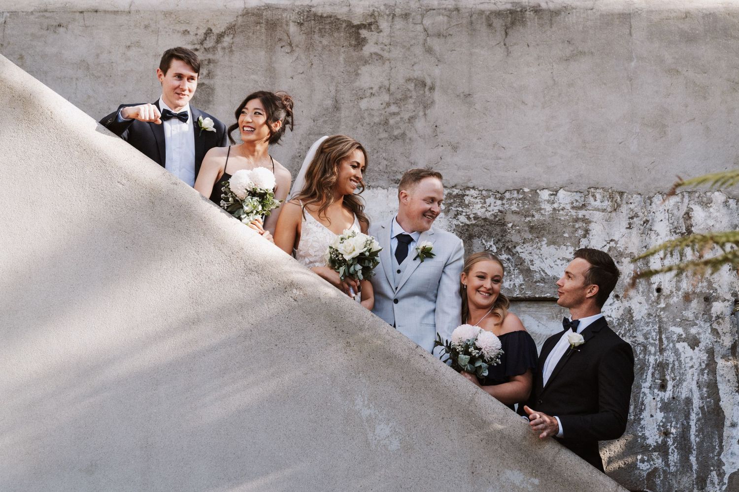 Wedding party posing on stone steps, bride holding bouquet, groom in light suit, guests in formal attire outdoors.
