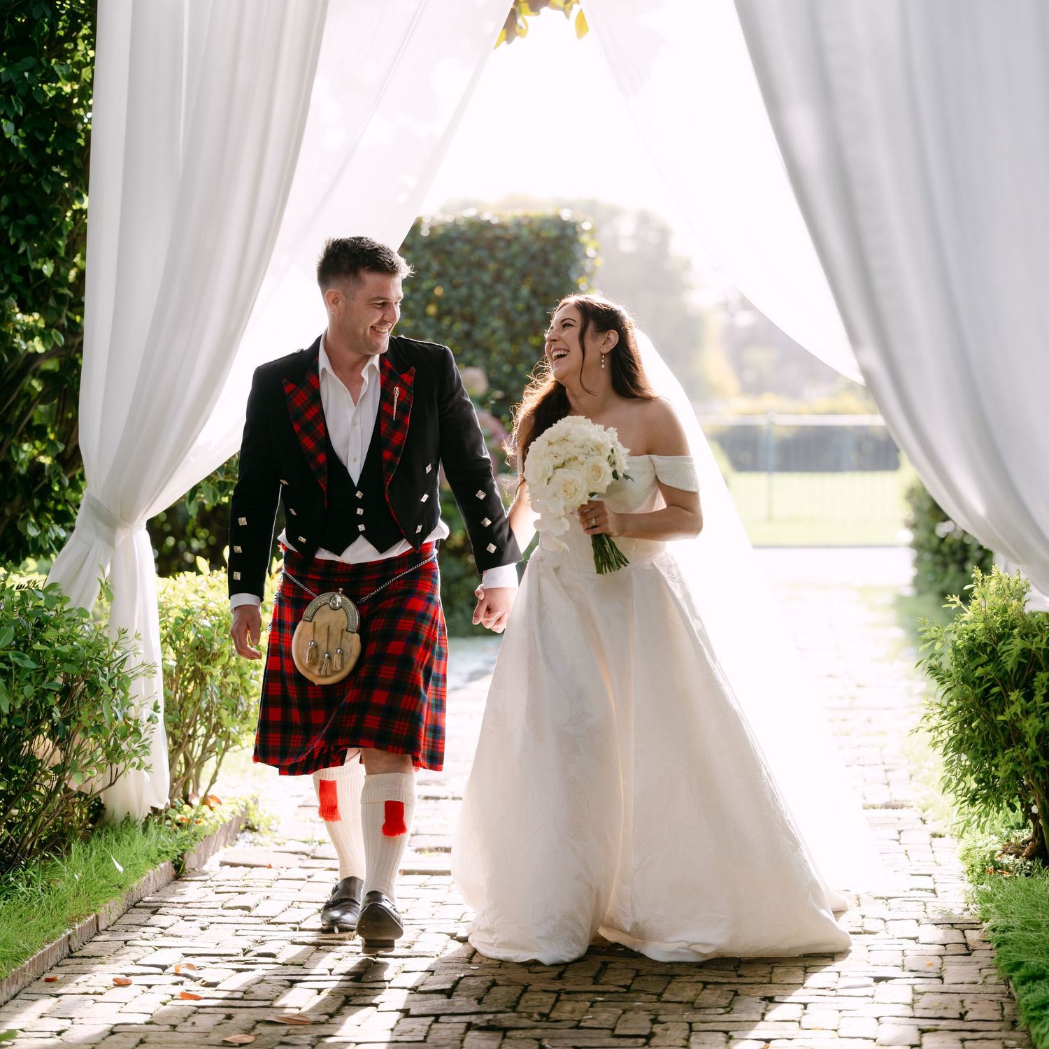 A groom in a red tartan kilt and bride in a white gown walk happily under white drapes at an outdoor wedding.