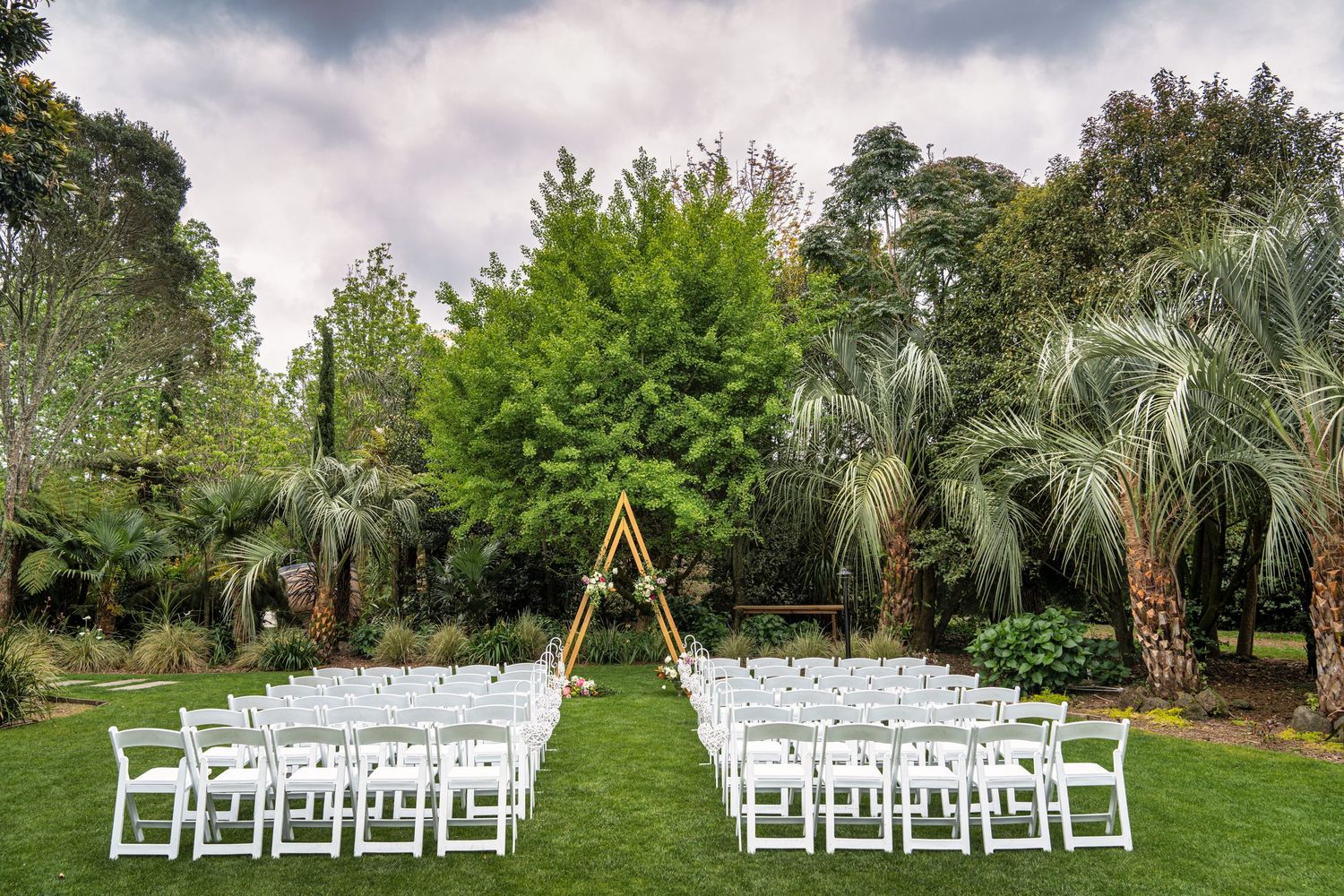 Outdoor wedding ceremony setup with white chairs, triangular arch, and lush green trees backdrop.