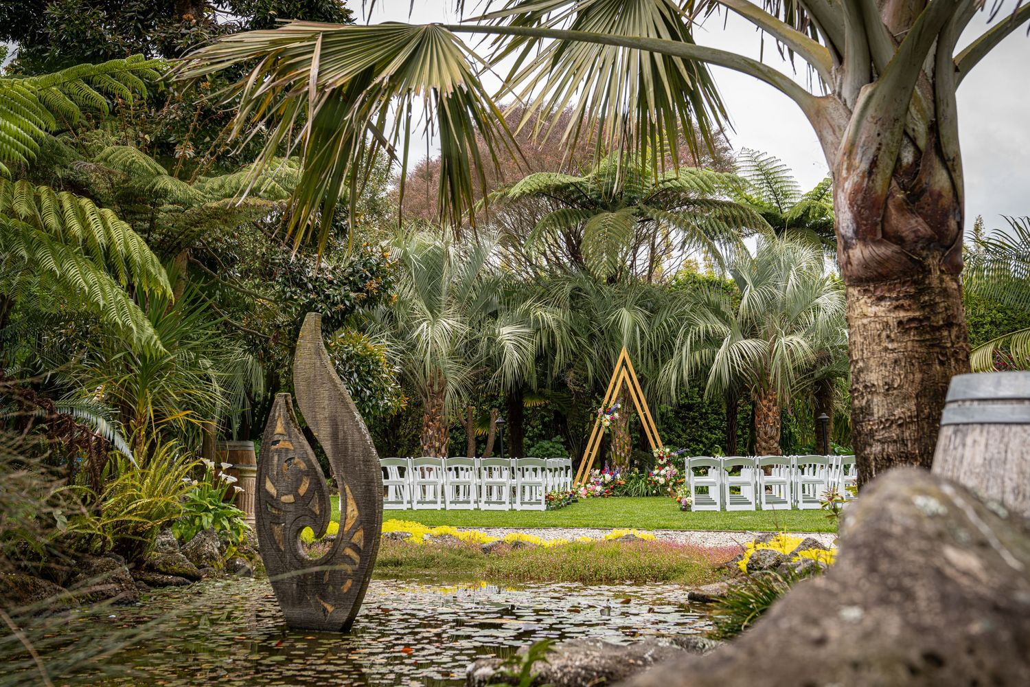 Tropical garden wedding ceremony setup with white chairs, palm trees, and wooden sculpture outdoor venue.