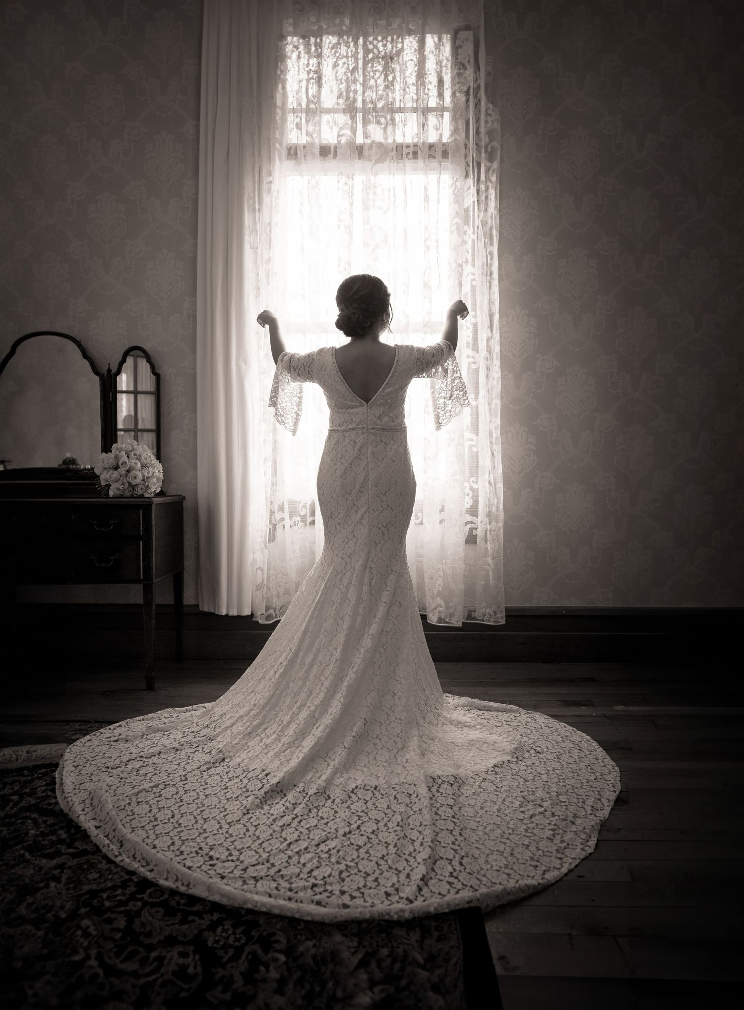 Black and white photo of a bride in a lace wedding gown with train, standing before a curtained window.