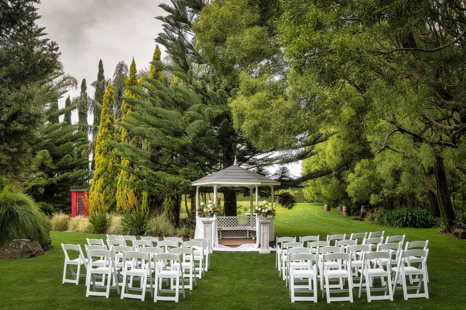 Outdoor wedding ceremony setup with white chairs arranged around a gazebo surrounded by lush green trees and gardens.