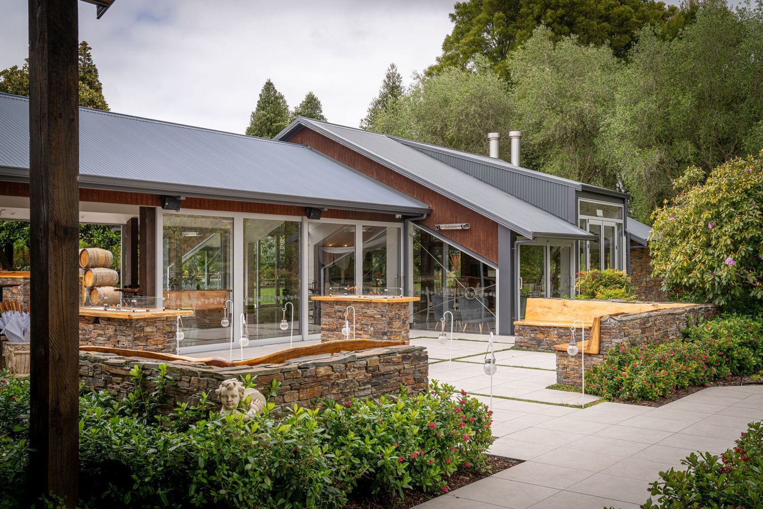 Modern stone and glass restaurant building surrounded by lush green gardens and trees on a sunny day.