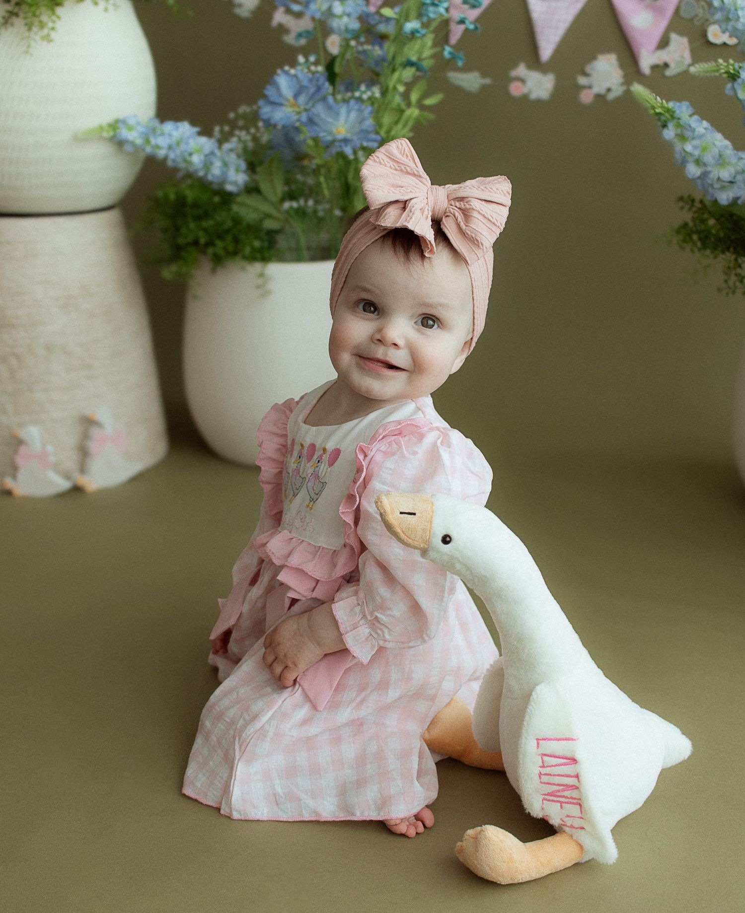Smiling baby girl in pink gingham dress and bow headband holding a white goose plush toy named Laine.