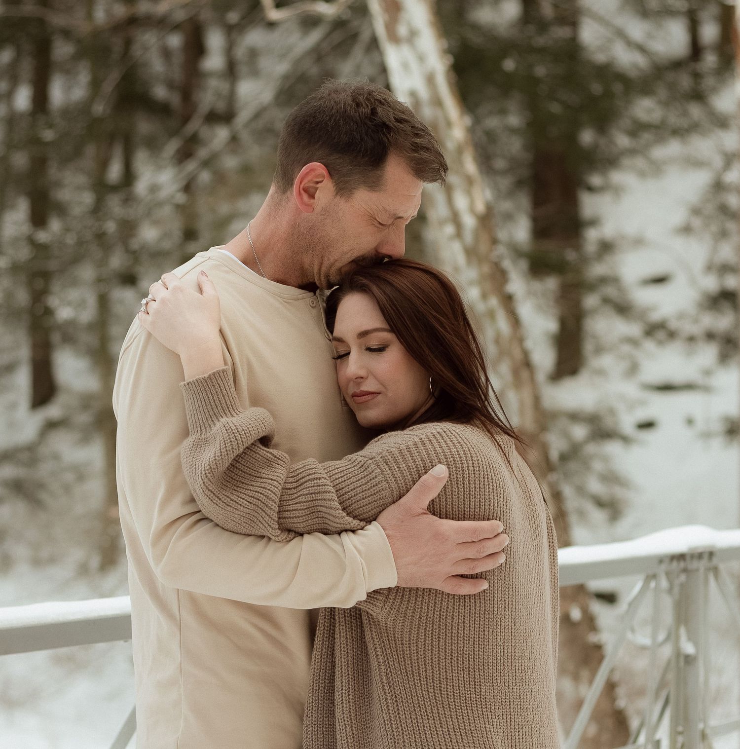 A couple in neutral tones embrace tenderly in a snowy winter forest setting.