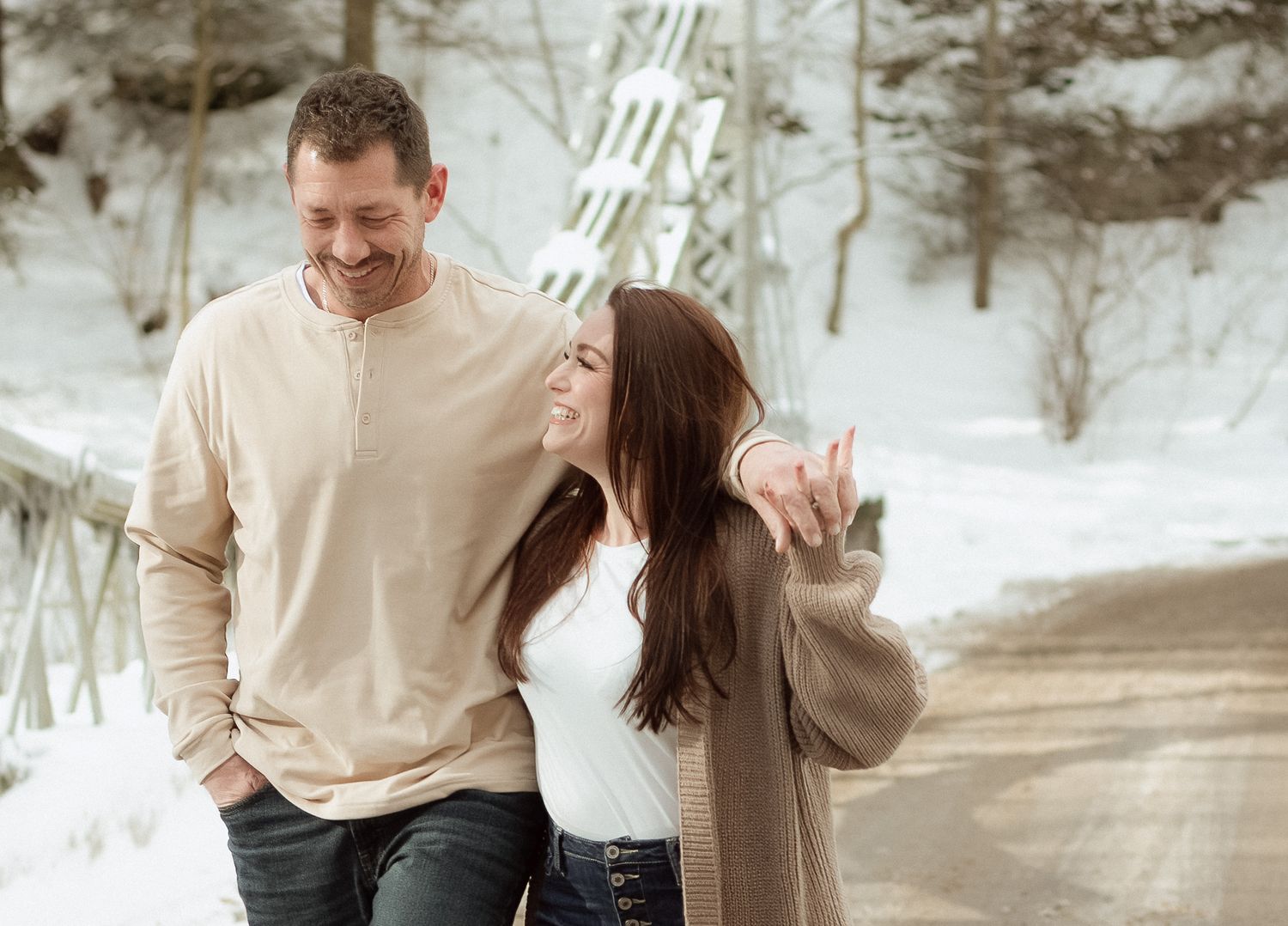 Couple laughing together outdoors in winter snow, wearing casual warm clothing near a bridge.